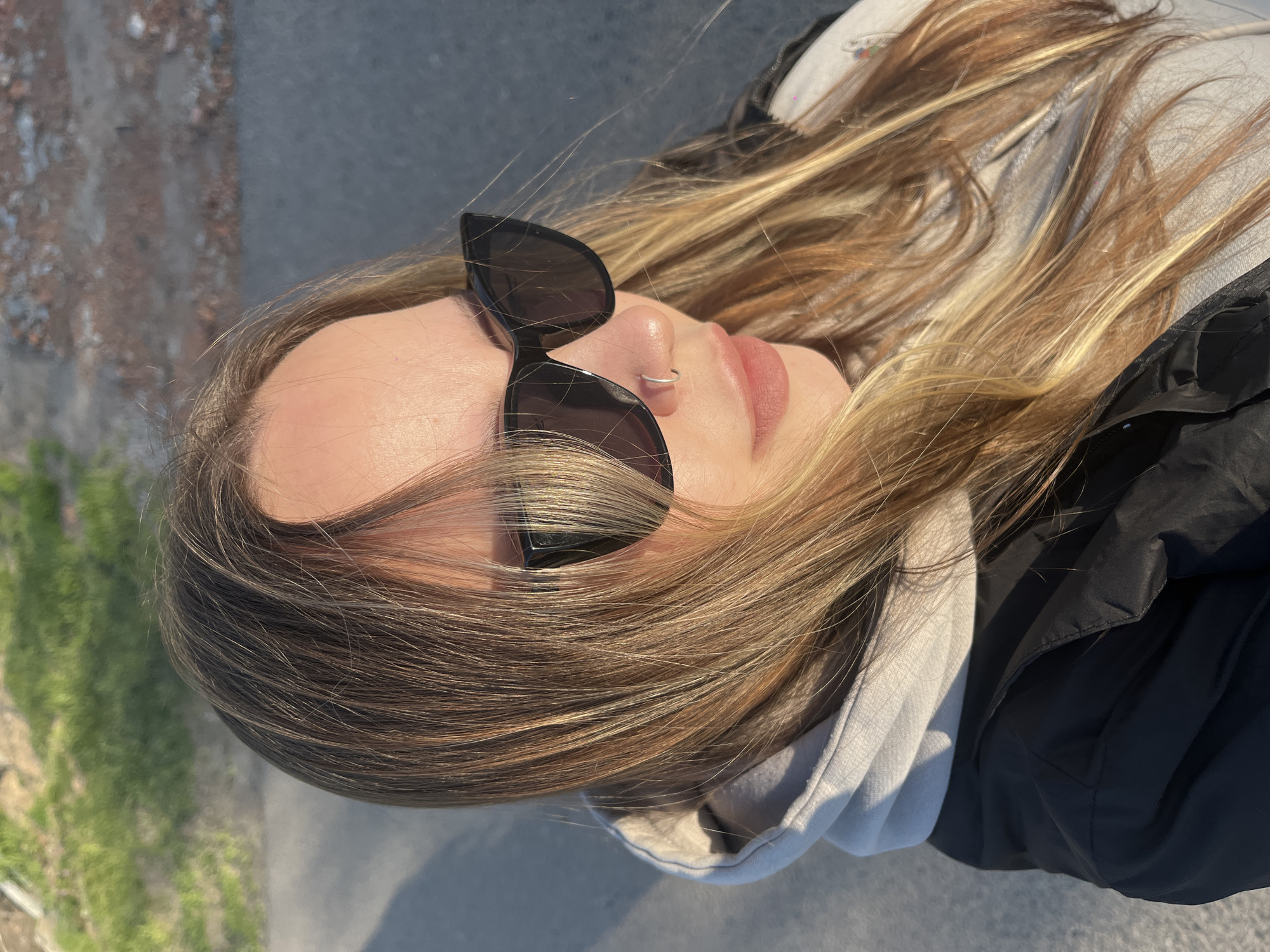 The image displays a close-up, upward-angled portrait of a young woman lying down outdoors, her head tilted slightly. She is seen from the forehead down to just below her chin. Her eyes are obscured by dark, cat-eye style sunglasses, and she has a small silver hoop piercing in her nose. Her lips are naturally pink. Her hair is long, a blend of light brown and blonde highlights, flowing around her head.
She is dressed in casual outerwear, including a light grey hooded sweatshirt worn beneath a dark, likely black, jacket with a textured, possibly puffy, material. The bright sunlight suggests a clear, sunny day, casting soft shadows. The background is out of focus, indicating an outdoor setting. Visible elements in the blurred background include dark, smooth surfaces resembling pavement or a road, along with some lighter, textured areas that could be gravel, stones, or a distant building. A hint of green vegetation, possibly grass, is also discernible. The overall atmosphere is relaxed, enjoying the sunny weather. There is no visible text in the image. The location context is Novopokrovskaya, Russia.