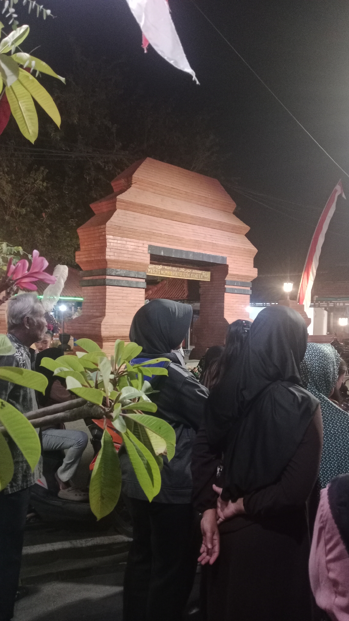 The image depicts a nighttime scene in front of a brick building with an arched entrance, identified by Arabic script and the text "MASJID NURHIDAYATULKOH KUNINGAN" in Latin script, suggesting it is a mosque. Several people are present, mostly wearing dark clothing and headscarves, indicating a likely religious or cultural gathering. The lighting is artificial, casting shadows and creating a somewhat dim atmosphere. In the foreground, there are some leafy branches obscuring the view. Indonesian flags are visible in the background. The scene appears to be taking place in a public area, possibly a street or a courtyard. The overall impression is one of a community event or gathering at dusk or night.