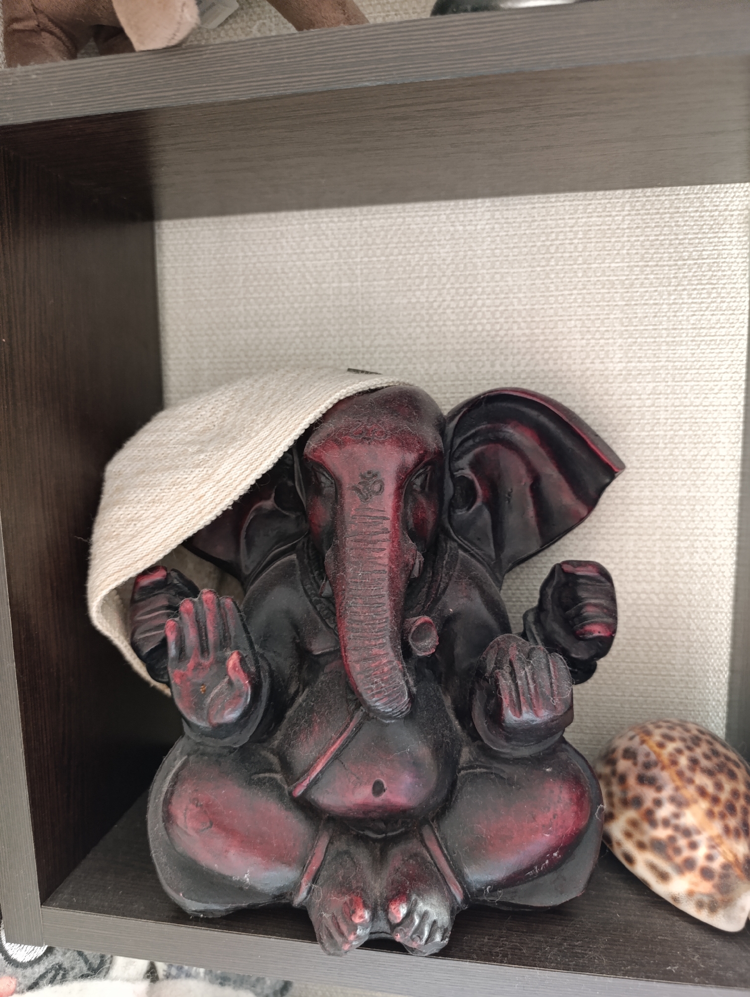 An ornate, dark red Ganesha statue is placed on a wooden shelf. The statue is covered from the top with a light-colored woven cloth. To the right of the statue is a spotted seashell. The background is a textured, light-colored wall. The scene is indoors, likely within a residential setting, and there is no information available to determine the specific location in Sertolovo, Russia.
