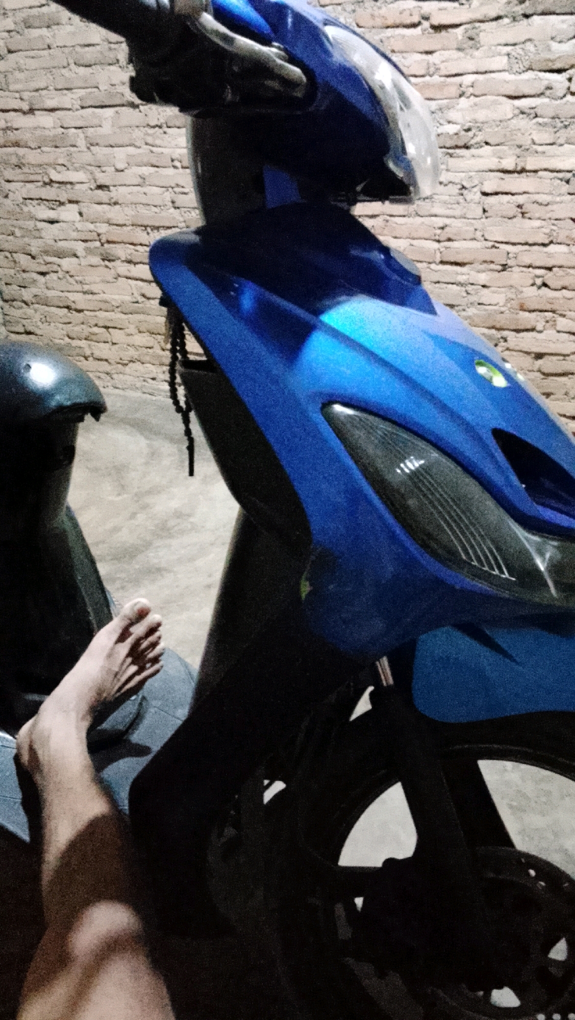 A close-up, vertical shot shows the front part of a bright blue motorcycle parked against a brick wall in Way Melan, Indonesia. A person's bare foot and lower leg are visible in the lower left of the frame, resting on what appears to be denim fabric. To the left of the foot, the dark silhouette of a motorcycle helmet is visible. A string of dark prayer beads hangs from the motorcycle's handlebars. The lighting suggests it is nighttime or dusk, casting shadows on the ground. The background is a textured brick wall, and the ground is a light-colored concrete surface.