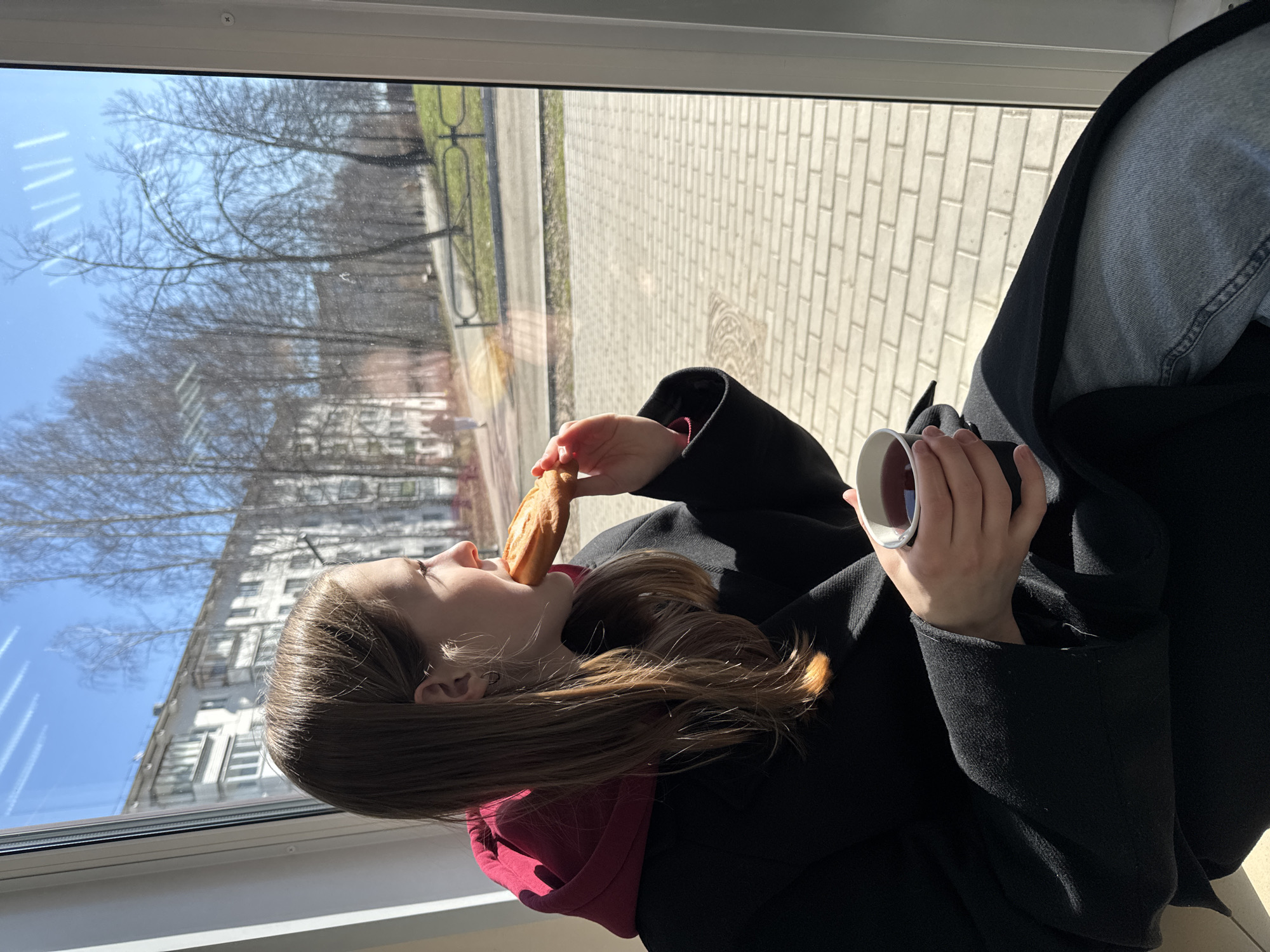 A young woman sits by a window, eating a pastry and holding a cup. She wears a black coat over a burgundy hoodie and blue jeans. Outside the window, there is a view of brick paving and trees, with a building visible in the distance. The location is likely Chaikovsky, Russia.