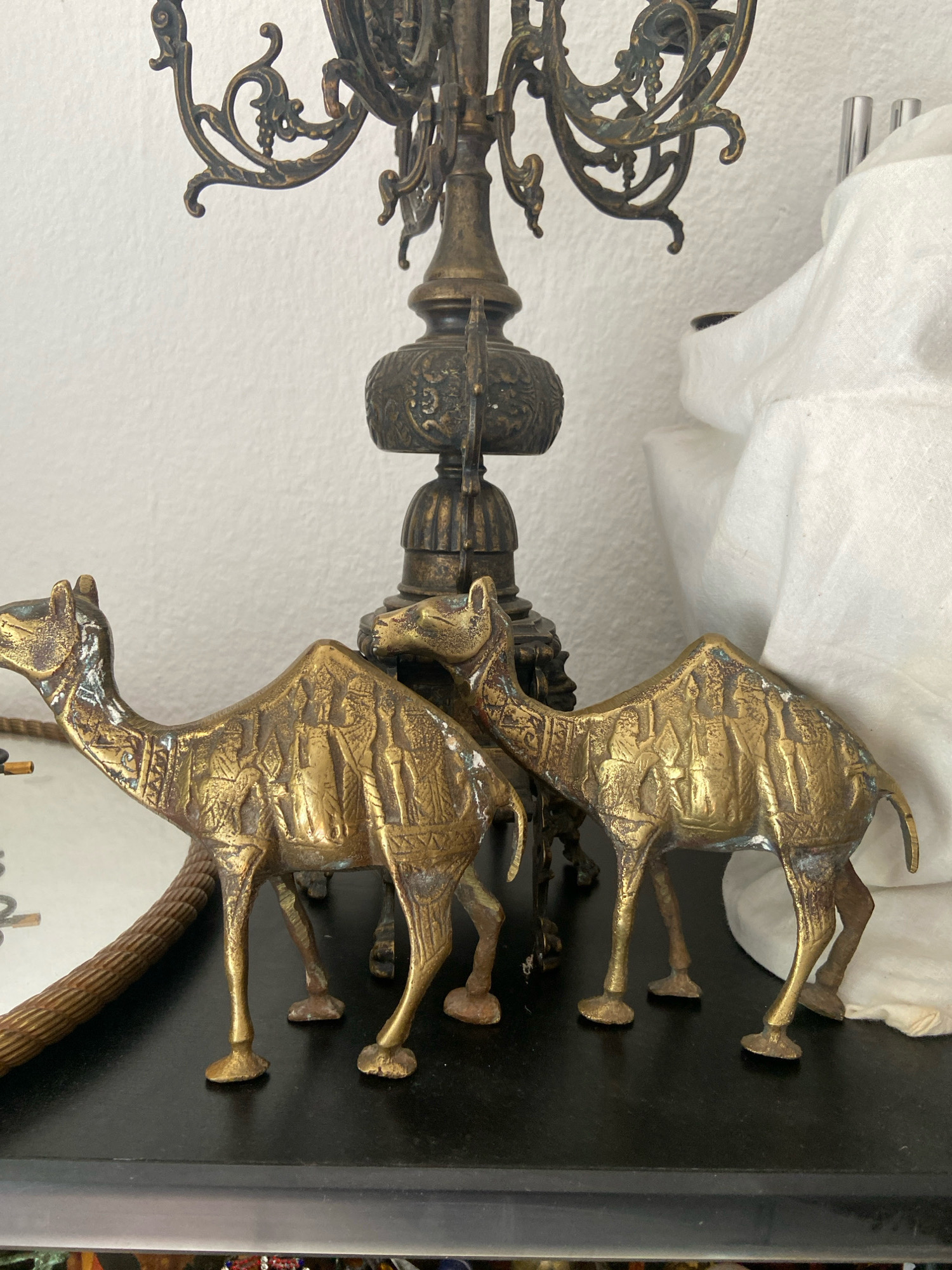 The image displays two brass camel figurines positioned on a dark, reflective surface. The camels are detailed, with the one on the left facing left and the one on the right facing right. Both figurines exhibit intricate carvings on their bodies, depicting what appear to be human figures in profile. Behind the camels, a portion of an ornate, antique-style brass candelabra is visible, suggesting an indoor setting. To the right of the candelabra, a white cloth is draped, partially obscuring the background. The lighting in the scene is soft, casting subtle shadows. The overall impression is that of decorative objects arranged in a domestic or display setting. There is no indication of the city of Kassel, Germany, in the image.