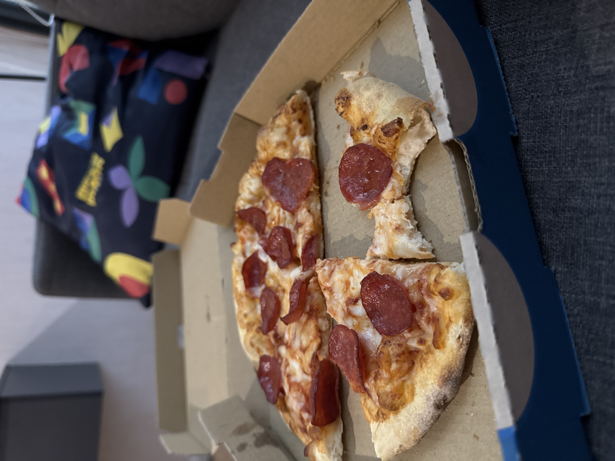 A partially eaten pepperoni pizza is in an open pizza box resting on a gray sofa. A patterned cushion is behind the box. The pizza box is blue and cardboard. The pizza has a thin crust and a layer of cheese and pepperoni.