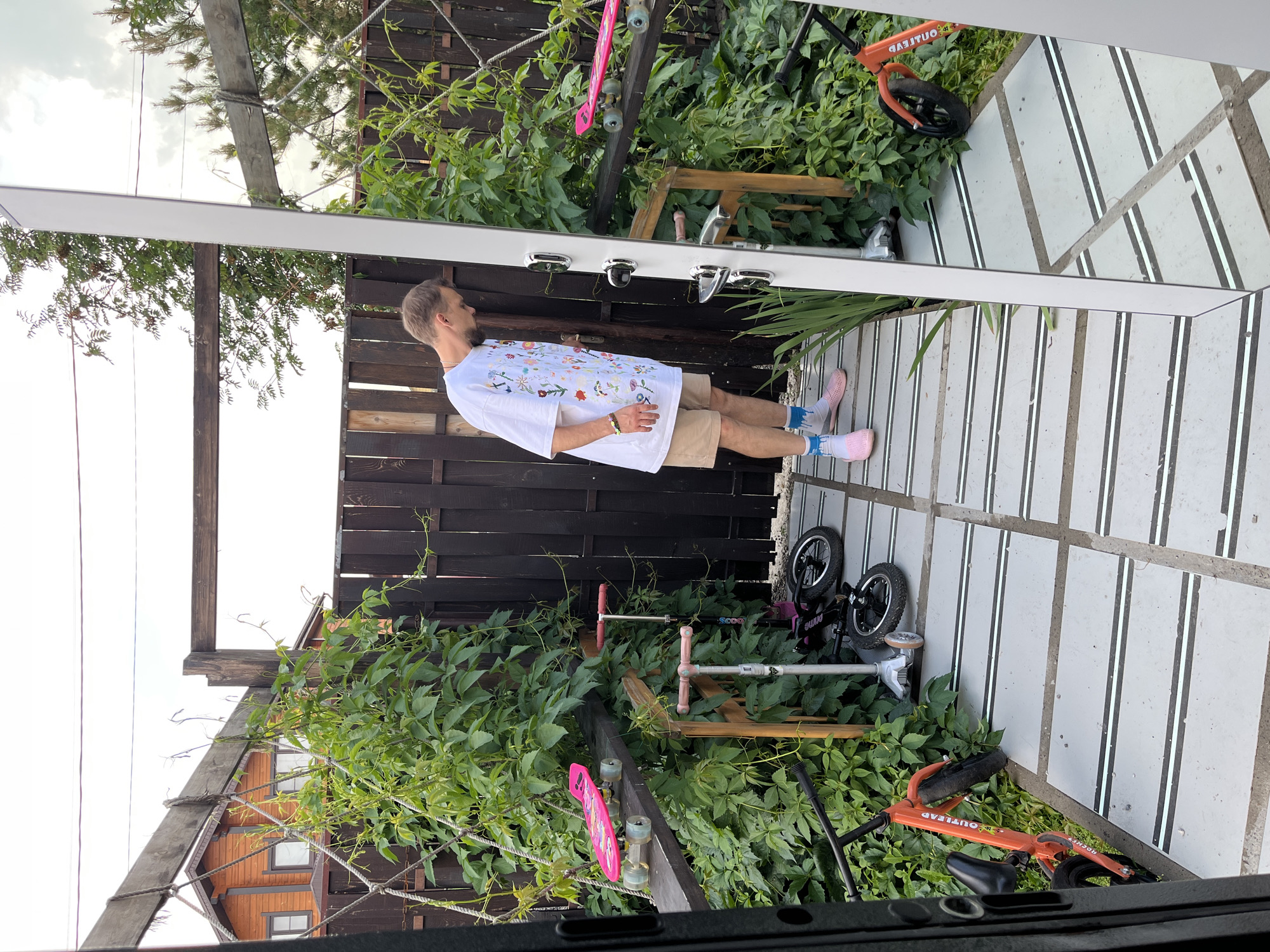 A man stands in an outdoor area in Sukhaia Balka, Russia, looking down. He is wearing a white t-shirt with a floral pattern, tan shorts, pink slip-on shoes, and white socks with blue stripes. The area appears to be a patio or walkway made of grey tiles with black and white stripes. To his left, a wooden fence and lush green vegetation are visible. Resting on a wooden beam above the vegetation are two pink skateboards. Several scooters are parked on the ground, including an orange one and two smaller silver and black ones. A large mirror is positioned to the right of the man, reflecting the scene. In the background, a wooden fence borders the area, and a two-story wooden house with windows is visible beyond it, partially obscured by trees. The sky is overcast.
