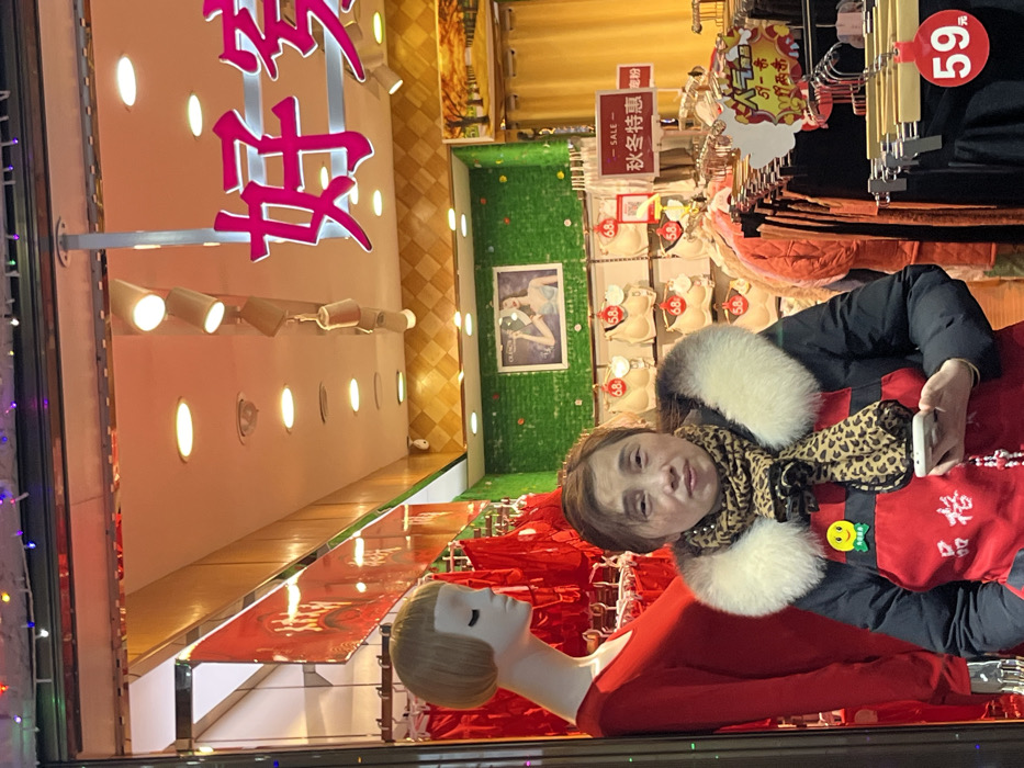 A retail storefront in Jiangjin, China, is depicted from an exterior perspective through a glass pane. The upper edge of the frame shows multicolored string lights reflected on the glass.
In the mid-ground, a female individual of East Asian descent stands, looking directly at the camera. She wears a black puffer coat with a prominent light-colored faux fur collar, a leopard print scarf, and a red apron or vest bearing white Chinese characters "品格" (pǐn gé) and a yellow smiley face pin on her left chest. She holds a white smartphone with a decorative beaded charm in her left hand.
To her left, a blonde female mannequin, visible from the chest up, wears a plain red long-sleeved garment. Its eyes are closed.
The store's interior features a brightly lit ceiling with multiple recessed lights, track lighting, and a dome-shaped security camera. A large pink neon sign displaying the Chinese characters "好家" (hǎo jiā) is prominent in the upper right. The walls are varied: a section with green textured material displays a framed image of a woman in lingerie, while an adjacent section has a diamond-patterned tan finish with a framed landscape artwork. Merchandise includes bras displayed on wall fixtures, each with a red tag indicating "68" as the price. A red sign reads "SALE" and "秋冬特惠" (qiū dōng tè huì, "Autumn Winter Special Offer"). Clothing racks with dark garments are visible in the lower right, marked with red "59" price tags.