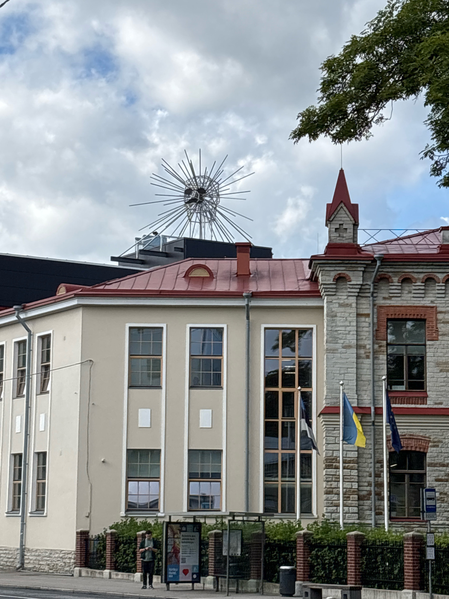The image shows a street view in Tallinn, Estonia, featuring two buildings. The building on the left is a light-colored structure with a red roof, featuring multiple windows. The building on the right is a more ornate stone building with a red roof and architectural details. A striking metal sculpture, resembling a sunburst or a spiky sphere, is visible above the rooftops. Several flags are displayed outside the stone building, including the Ukrainian flag and the European Union flag. A bus stop is located in the foreground, with a person standing nearby, looking at their phone. Green foliage and trees are also present in the scene, and the sky is cloudy.