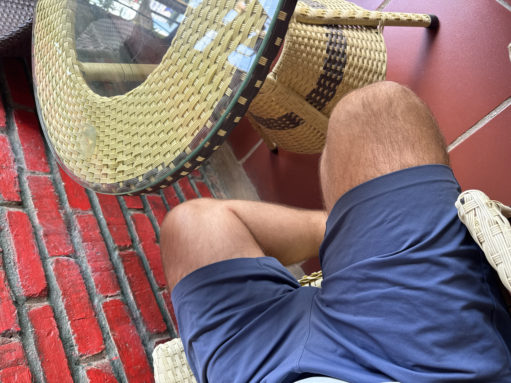 A person wearing blue shorts sits at a table in Đồng Văn Commune, Vietnam. The table has a glass top and a woven base. The floor is made of red bricks and brown tiles. A woven basket sits next to the table. The person's legs are visible from the knees up.