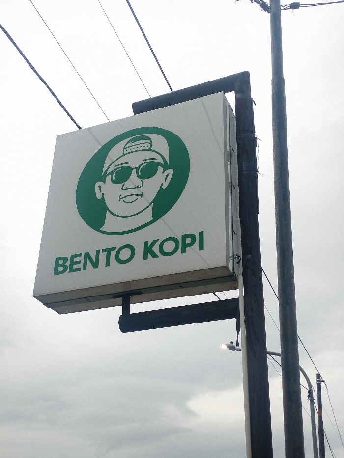 An outdoor, slightly low-angle shot captures a square white sign for "BENTO KOPI" against an overcast sky. The sign features a prominent green circular logo in its upper half. Within the green circle is a cartoon illustration of a person's face, depicted wearing a backward baseball cap and dark sunglasses, with a slight, cool smirk. The facial features are minimalist, with distinct ears and a chin.
Below the logo, the words "BENTO KOPI" are written in bold green sans-serif capital letters. The sign itself is illuminated from within, suggesting it's a lightbox, though the ambient light from the sky makes the internal light less distinct.
The sign is mounted on a dark, L-shaped metal frame which is attached to a sturdy, dark utility pole on the right side of the frame. Another thinner utility pole is visible behind the main one, and multiple dark electrical wires crisscross the upper portion of the frame and the sky, extending diagonally from the top left. A small, bright light fixture, possibly a streetlamp, is faintly visible in the lower right background, below the sign.
The sky is a uniform, light gray, indicating an overcast or cloudy day, with no direct sunlight. The overall scene suggests a commercial establishment in an urban or suburban area, common in places like Dramaga, Indonesia.