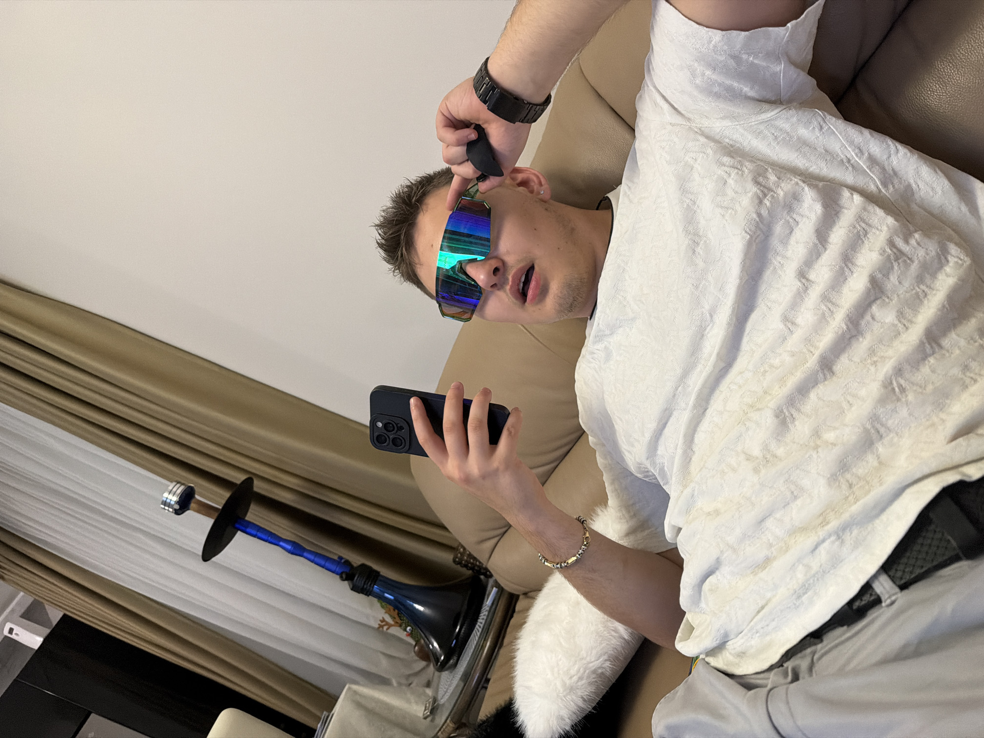 A person lies on a tan leather couch, holding a black smartphone in their left hand and adjusting a pair of iridescent sunglasses with their right. The person is wearing a white textured shirt, grey pants, and a black belt. A black watch is on their right wrist, and a gold bracelet is on their left. To the left, a blue and black hookah sits on a glass table covered with a white furry rug. The background features a wall partly covered by tan curtains. The location is Unknown, Unknown.