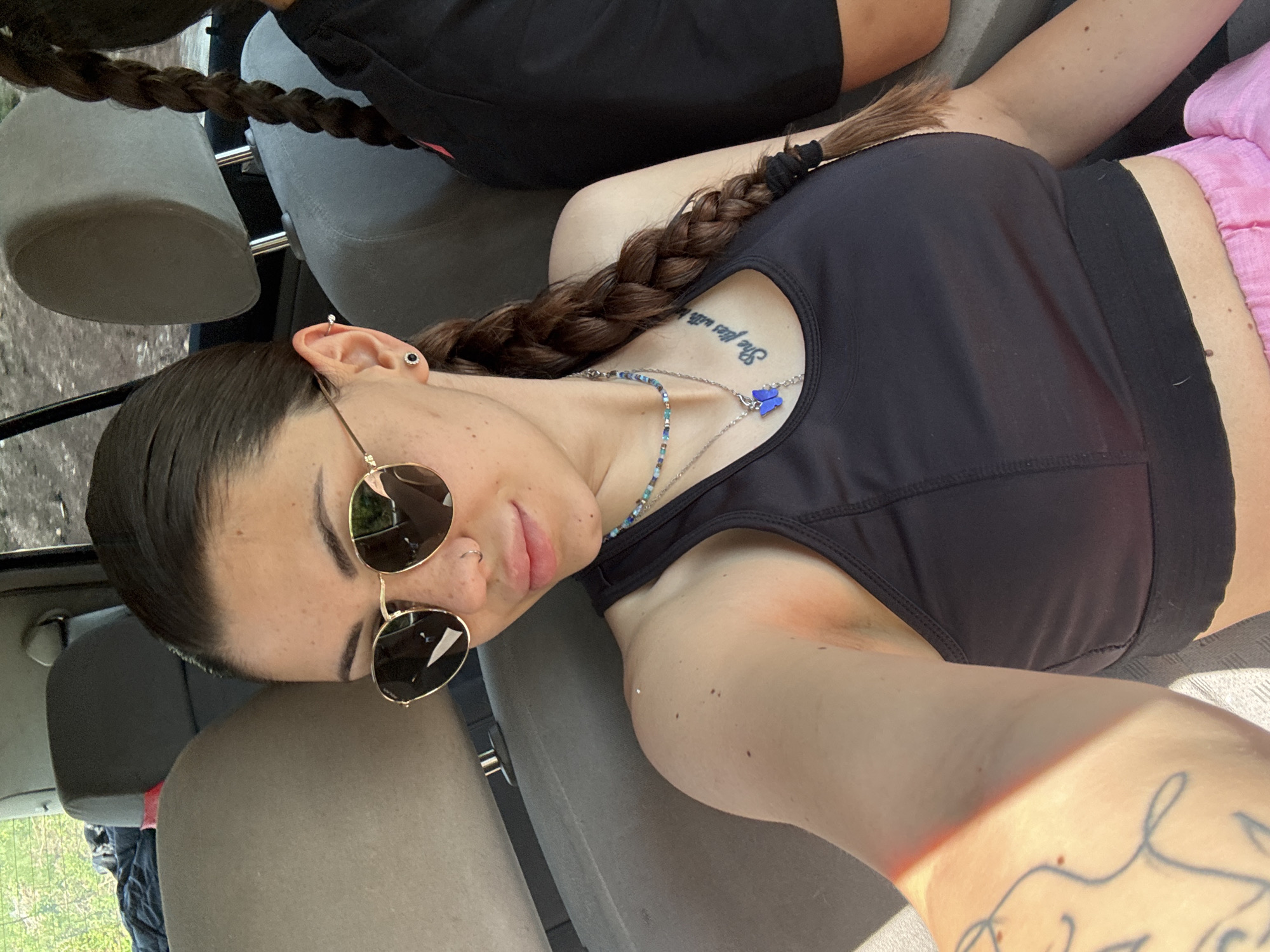 A young woman is captured in a close-up selfie, lying down in what appears to be the passenger seat of a car, with the image rotated counter-clockwise. She has dark, braided hair that extends over her shoulder and chest. She is wearing a black sports bra and partially visible pink shorts. Her face is turned slightly towards the camera, adorned with dark, round sunglasses and multiple piercings in her ear (helix and lobe) and nose. A relaxed, subtle smile plays on her lips.
Around her neck, she wears two necklaces: one with small colorful beads and another silver chain featuring a distinctive blue butterfly pendant. A tattoo is visible on her upper chest, reading "She flies with...", with the rest of the text obscured by her braid. Another tattoo can be partially seen on her right arm.
Bright sunlight streams into the car, casting strong highlights on her skin and creating distinct shadows, suggesting it is daytime and sunny. In the background, out of the car window, green foliage is visible, indicating an outdoor setting, possibly rural or natural. Part of another person's lower body, dressed in black, is visible in the background, suggesting two people are in the car. The scene takes place in Belopoptsi, Bulgaria.