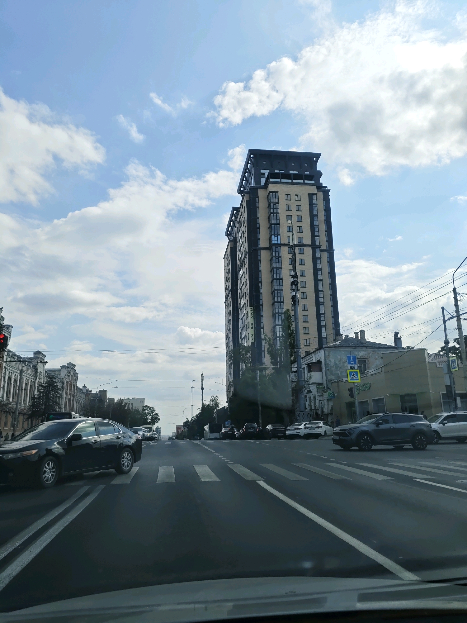The image depicts a street scene in Rostov-on-Don, Russia, on a partly cloudy day. A tall, modern apartment building with a dark facade and light-colored sections dominates the right side of the frame. In the foreground, a dark-colored sedan is driving on a multi-lane road with white road markings, including a crosswalk. Other vehicles are visible on the road, including a gray SUV crossing the crosswalk, and several other cars in the distance. Along the left side of the street, older buildings with ornate architectural details are visible. Traffic lights and power lines are present. The sky is blue with scattered white clouds. The overall impression is of a busy urban environment.