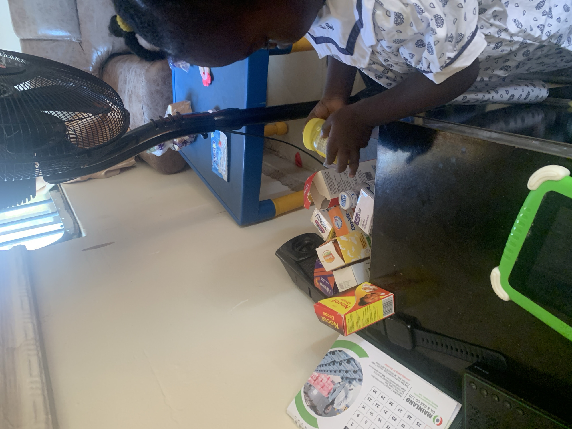 The image captures a child in Gura Top, Nigeria, interacting with a collection of medicines on a dark surface. The child, wearing a patterned white dress, is holding a yellow container, likely medicine, in their hands. Several medicine boxes are visible, including one labeled "Nocof Drops," another with "Em-Vit," and one with "Abidec." A calendar with the "MAINLAND OIL & GAS CO. LTD" logo is also present. The scene appears to be indoors, with a fan and a blue table with yellow legs in the background. The lighting suggests it's daytime, with light coming from a window.