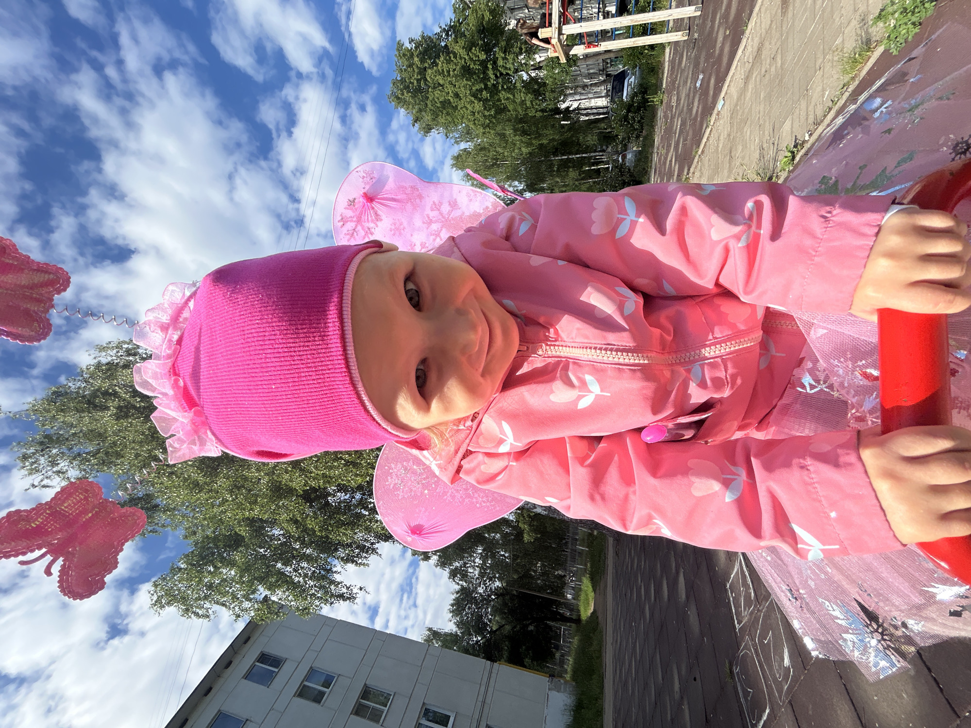 This media file presents a photograph rotated 90 degrees clockwise, capturing a young child in a vibrant pink fairy costume. The child, likely a toddler, is centered in the lower-left portion of the frame, gazing directly at the viewer with a gentle, open-mouthed smile. They wear a bright pink knit beanie, a pink jacket embellished with light-colored cherry or flower patterns, and a sheer, glittery pink tutu, complemented by a pair of translucent pink fairy wings on their back. Their hands firmly grasp a red bar, suggesting they are on a playground structure.
The background on the left and upper sections of the rotated image features a vivid blue sky dotted with scattered white clouds. Beneath the sky, the dense green foliage of deciduous trees is visible, indicating a warm season. A pink butterfly-shaped balloon is partially seen on the far left, connected by a coiled wire.
To the right, the scene transitions to an urban environment. A paved ground area displays faint chalk drawings, possibly numbers or symbols. Beyond this, multi-story grey apartment buildings with numerous windows rise amidst more trees and distant infrastructure. The image is brightly lit by natural daylight, highlighting the sunny weather. The overall setting points to an outdoor play area or park in Chaikovsky, Russia, where the child is either engaged in play or posing for the camera.