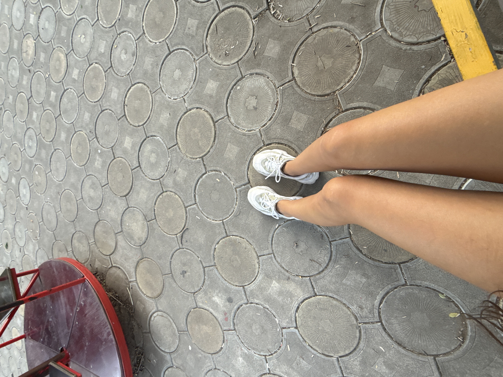 The image depicts a first-person perspective of a person sitting on a yellow bench, with their legs extended forward. The person is wearing white athletic shoes. The ground is paved with circular concrete blocks arranged in a grid pattern. To the left, a portion of a red merry-go-round is visible. The scene appears to be an outdoor public space, likely a playground or park area, in the city of Basarabeasca, Moldova. The lighting suggests it is daytime.