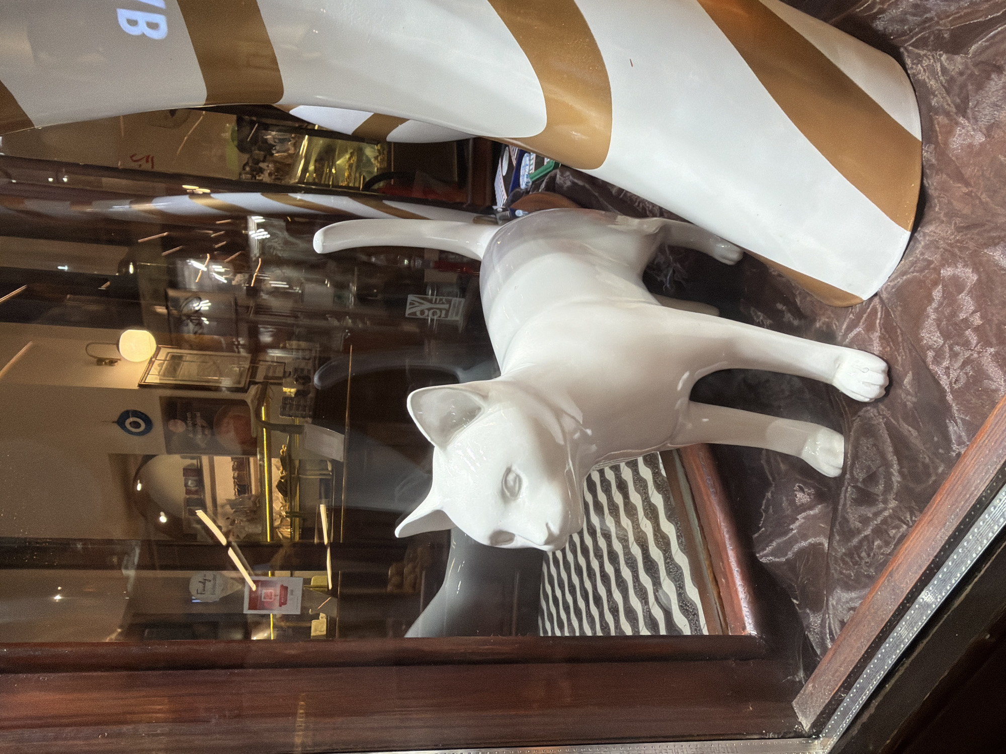 A white ceramic cat statue stands in a shop window in Kadıköy, Turkey. The cat is facing to the left, with its head slightly turned towards the viewer. It is standing on a dark brown, textured fabric that drapes around the base of a large, white, cylindrical object decorated with gold stripes. Behind the cat, a checkered black and white pattern is visible on the floor. The shop window reflects the interior of the shop, showing displays of baked goods and decorative items, including a blue evil eye charm hanging on the wall. To the right of the cat, a portion of the gold-striped cylindrical object is visible, appearing to be a decorative element. The overall scene suggests a display in a bakery or confectionery shop.