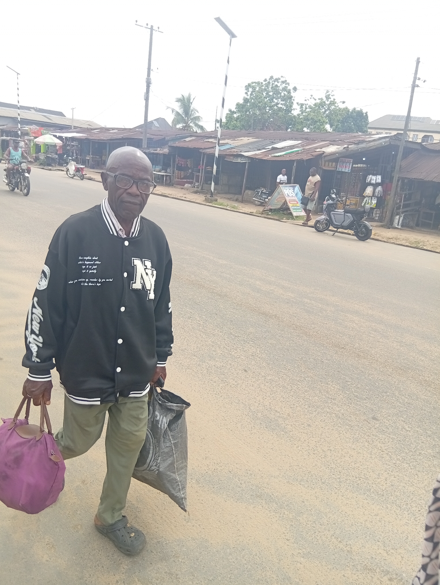 The image shows an elderly man in Aba, Nigeria, walking down a street lined with small shops. He is wearing a black varsity jacket with "New York" written on the sleeve and an "N" on the chest, khaki pants, and Crocs. He carries a purple tote bag in one hand and a black plastic bag in the other. The street appears to be paved, and there are motorcycles parked along the side. The shops have corrugated metal roofs and are open-fronted, with merchandise displayed. There are pedestrians and motorcycles on the street. Overhead power lines are visible.
