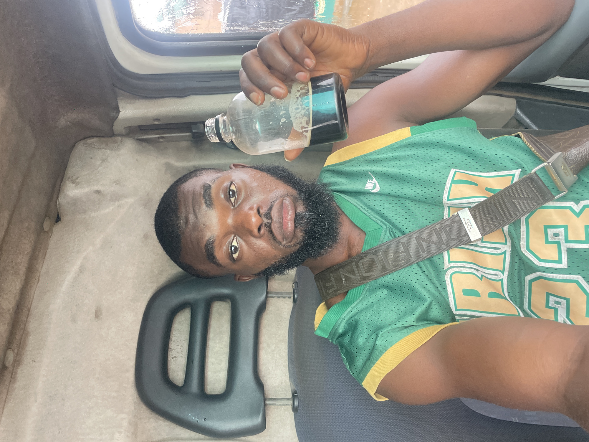 A man sits in the driver's seat of a vehicle, holding up a clear glass bottle filled with a dark liquid. He is wearing a green basketball jersey with yellow trim and a dark strap across his chest. The interior of the vehicle appears to be a truck cabin, with a black headrest visible behind him. The man has dark skin, a beard, and is looking directly at the camera with a neutral expression. The background outside the vehicle is blurred, but appears to be outdoors with some greenery and possibly buildings. The image is taken from a slightly low angle. It is not possible to determine the location in the city of Jos, Nigeria from the provided image.