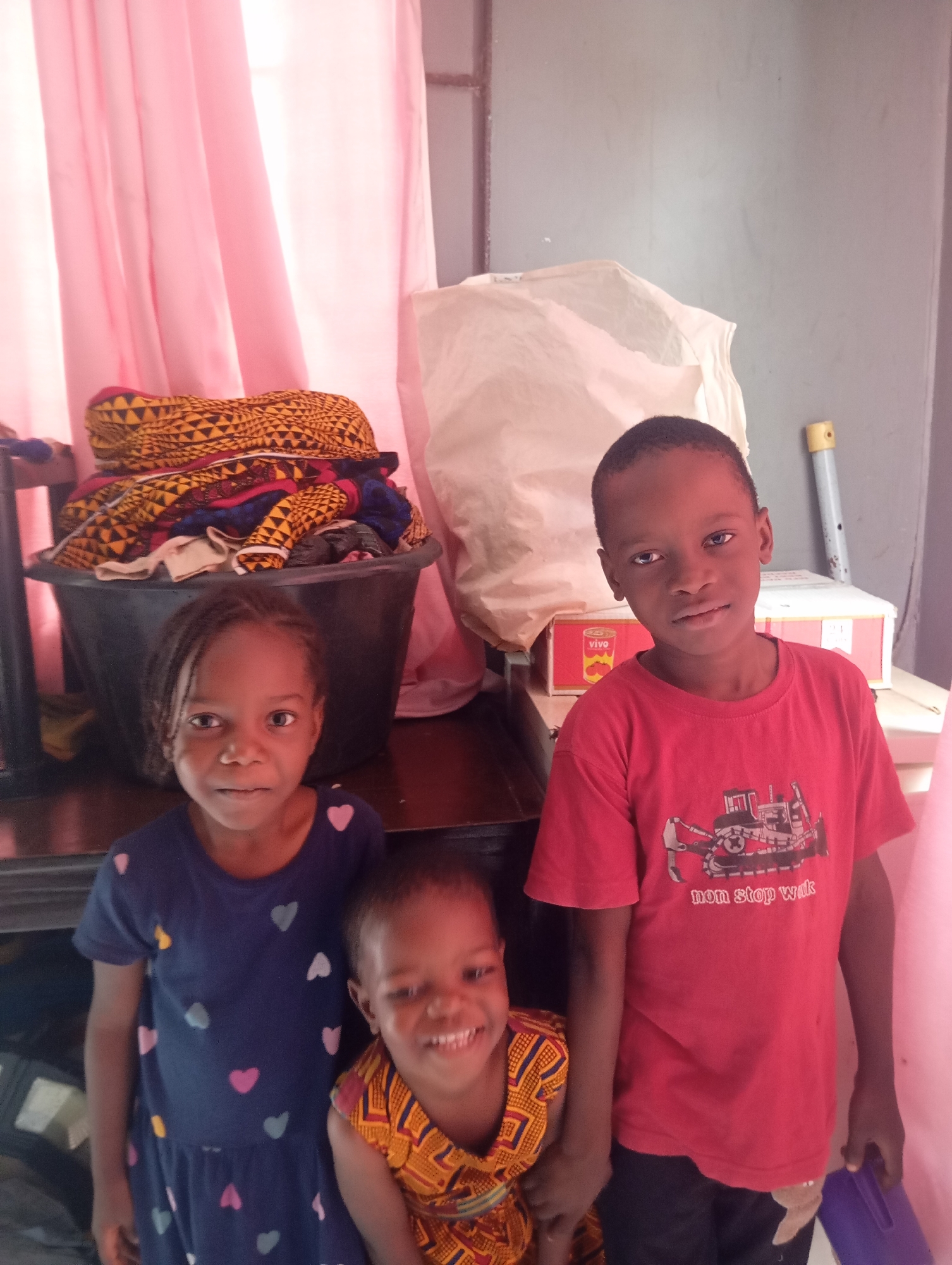 In Nsugpe, Nigeria, three children pose for a photo indoors. On the left, a young girl with braided hair wears a navy blue dress adorned with colorful heart patterns. To her right, a younger child smiles broadly, dressed in a vibrantly patterned yellow and red outfit. Standing on the far right is a boy wearing a red t-shirt with a graphic of a bulldozer and the text "non stop wak." Behind the children, a large black tub filled with colorful fabrics is visible, next to a crumpled white sheet or bag. Pink curtains hang in the background, and a portion of a gray wall is also present. A yellow and blue marker and a white box are on a shelf to the right of the boy.