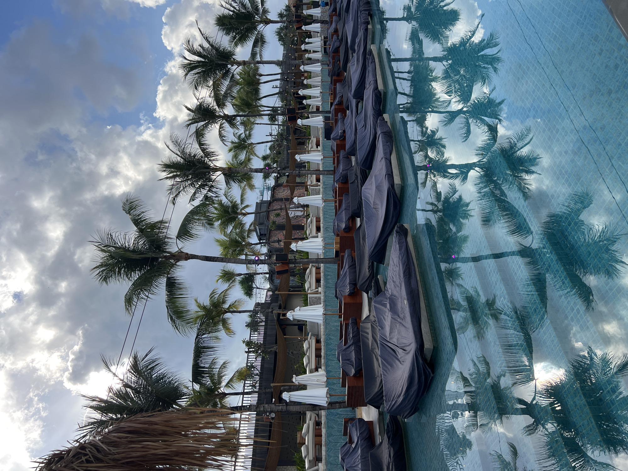 This is an outdoor scene with a swimming pool in the foreground, reflecting palm trees and clouds. In the background, there are many lounge chairs, some covered with dark fabric, and white umbrellas. Behind the lounge chairs, there is a building with a fence and more palm trees. The sky is partly cloudy with sunlight peeking through. The image appears to be taken at a resort or a beach club in North Kuta, Indonesia, given the prevalence of palm trees and the tropical setting. There are no people visible in the image. The water is clear and tiled with blue mosaic tiles.