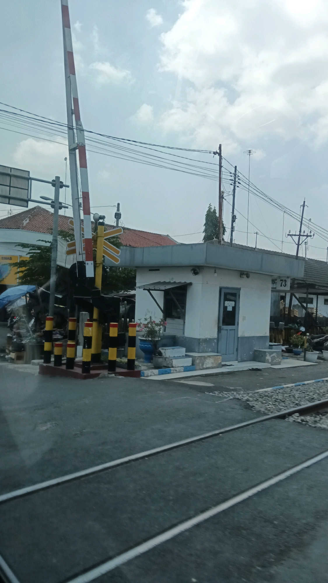 This image depicts a railroad crossing in Sragen, Indonesia, likely near the Sragen train station, given the presence of a small guardhouse. The scene is captured from a slightly elevated perspective, possibly from a moving vehicle.
In the foreground, a set of railroad tracks runs across a wet asphalt road. The tracks themselves are flanked by gravel.
To the left, a prominent railroad crossing gate is visible, with red and white striped poles angled upwards. Below the gate, a yellow and black striped post with reflective elements stands as a warning. Several smaller black and yellow striped bollards are arranged in a line.
A small, white, one-story guardhouse with a light blue door and window frames is situated to the right of the crossing gate. The guardhouse has a simple metal awning over the window and appears to be the operational center for the crossing. The number "73" is visible on the right side of the guardhouse. A potted plant with pink flowers sits near the guardhouse entrance.
In the background, a red-tiled roof of a building is visible, suggesting it might be part of the train station complex or nearby infrastructure. Electrical wires and utility poles crisscross the sky, which is overcast with scattered clouds. Some greenery, including trees, is present behind the crossing gate and guardhouse. The overall impression is of an active, functional railroad crossing on a somewhat cloudy day.