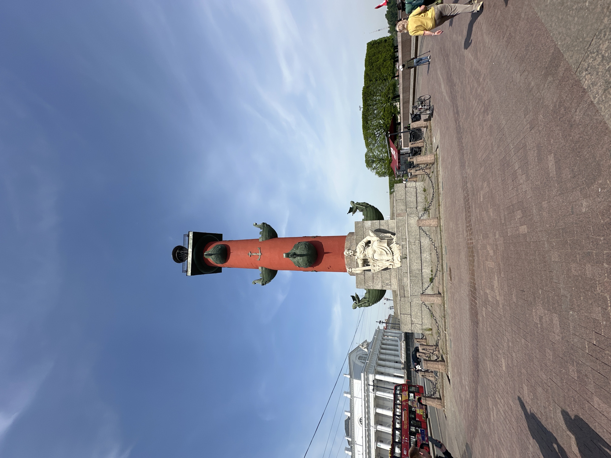 The image depicts the Rostral Columns of St. Petersburg, Russia. These are two rostral columns standing on the Strelka (eastern tip) of Vasilyevsky Island. The structures are red columns adorned with ship prows and are topped with beacons. In the background, a clear blue sky is visible with wisps of clouds. The surrounding area includes a paved square, with a view of buildings and buses in the vicinity, indicating a public space or square. People are walking in the upper right part of the image.