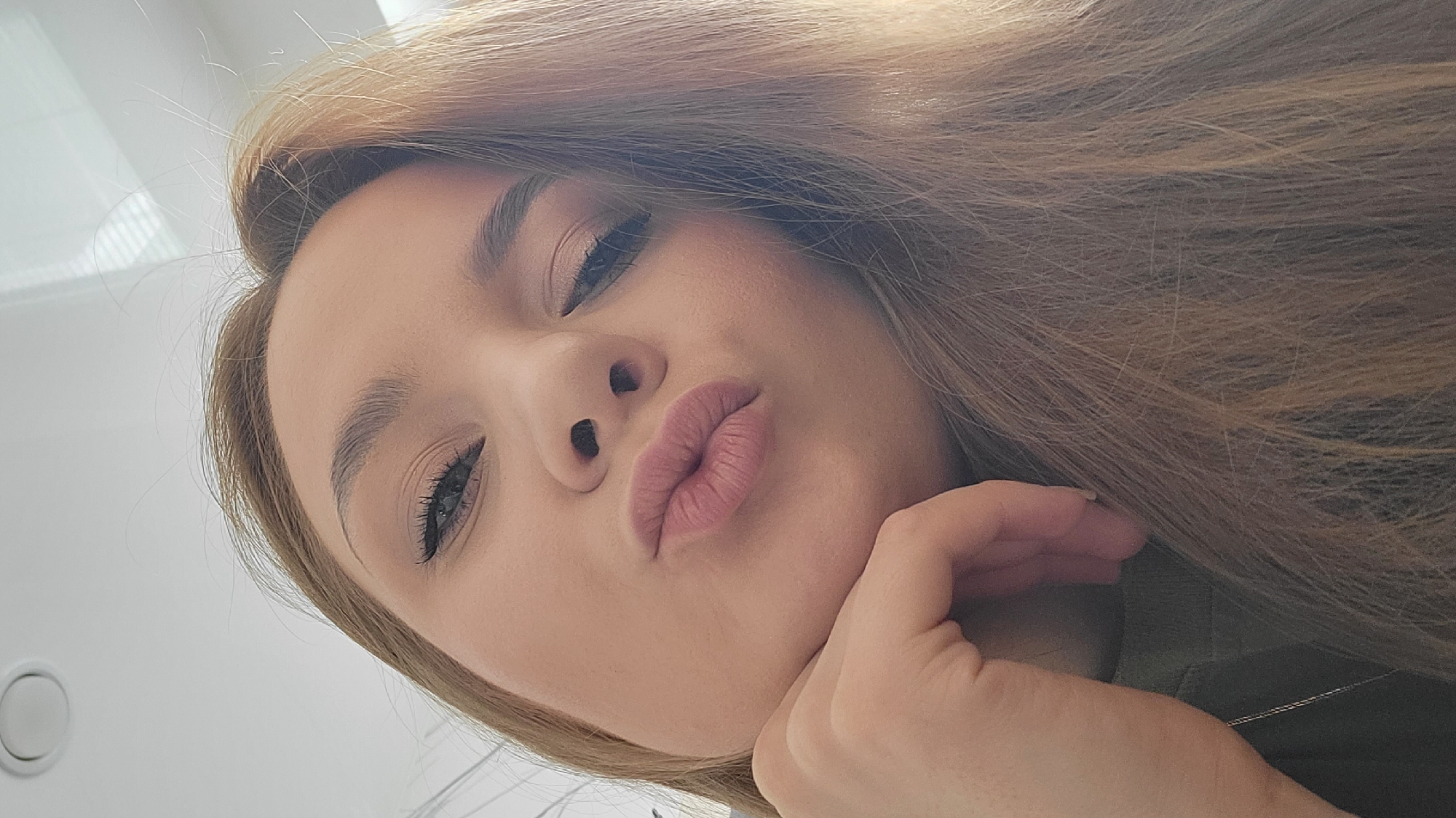 The image shows a woman with long, light brown hair, wearing makeup, including eyeshadow and mascara, making a kissing face towards the camera. She is resting her chin on her hand. A small part of what appears to be a green shirt is visible. The background is out of focus and predominantly white, revealing a light fixture or fitting on the upper left corner. The image was not taken in Chaikovsky, Russia, or any other identifiable specific location.