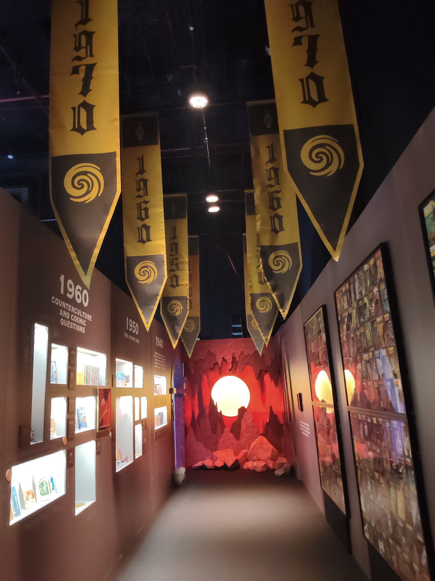 The image depicts a dimly lit exhibition hall with a walkway. Yellow banners with black text and circular symbols hang from the ceiling. The text on the banners reads "1970", "1960", "1950", and "1940" on successive banners. The walkway is lined with display cases on the left, exhibiting books and artifacts. On the right wall, framed artwork is displayed. At the end of the walkway, a stylized rock formation with a large circular light in the center is visible, resembling a sunset or a celestial body. The environment appears to be indoors, likely a museum or exhibition space. There are no people visible in the image. The overall impression is that of a historical exhibition, possibly focusing on different decades or eras. The location is not specified in the image.