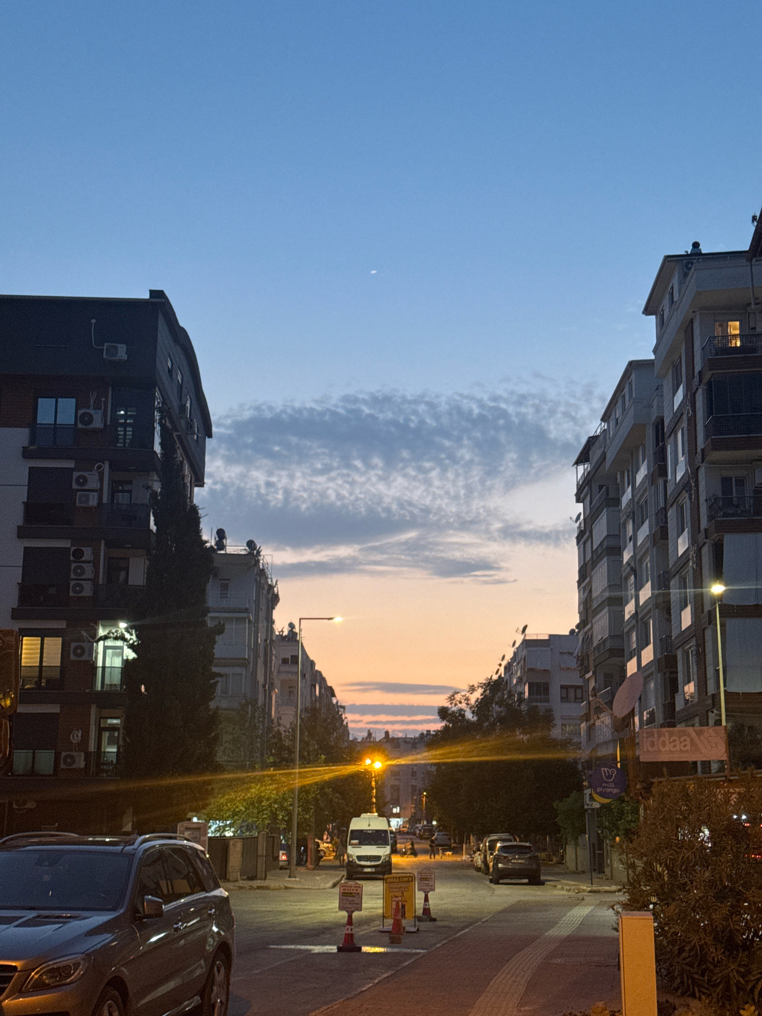 This image depicts a street scene in Antalya, Turkey, at dusk. Tall apartment buildings line both sides of the street. On the left, a dark-colored building has multiple air conditioning units visible on its facade. On the right, a lighter-colored building features balconies and illuminated windows. The sky is a gradient of blue and orange, with wispy clouds. A slender crescent moon is visible in the upper part of the sky. A street lamp with a bright yellow light illuminates the street, casting long shadows. Several cars are parked or driving on the street. One car in the foreground is a dark SUV. A white van is parked further down the street. Traffic cones and warning signs are placed along the side of the road. The overall impression is a calm evening in a residential urban area.
