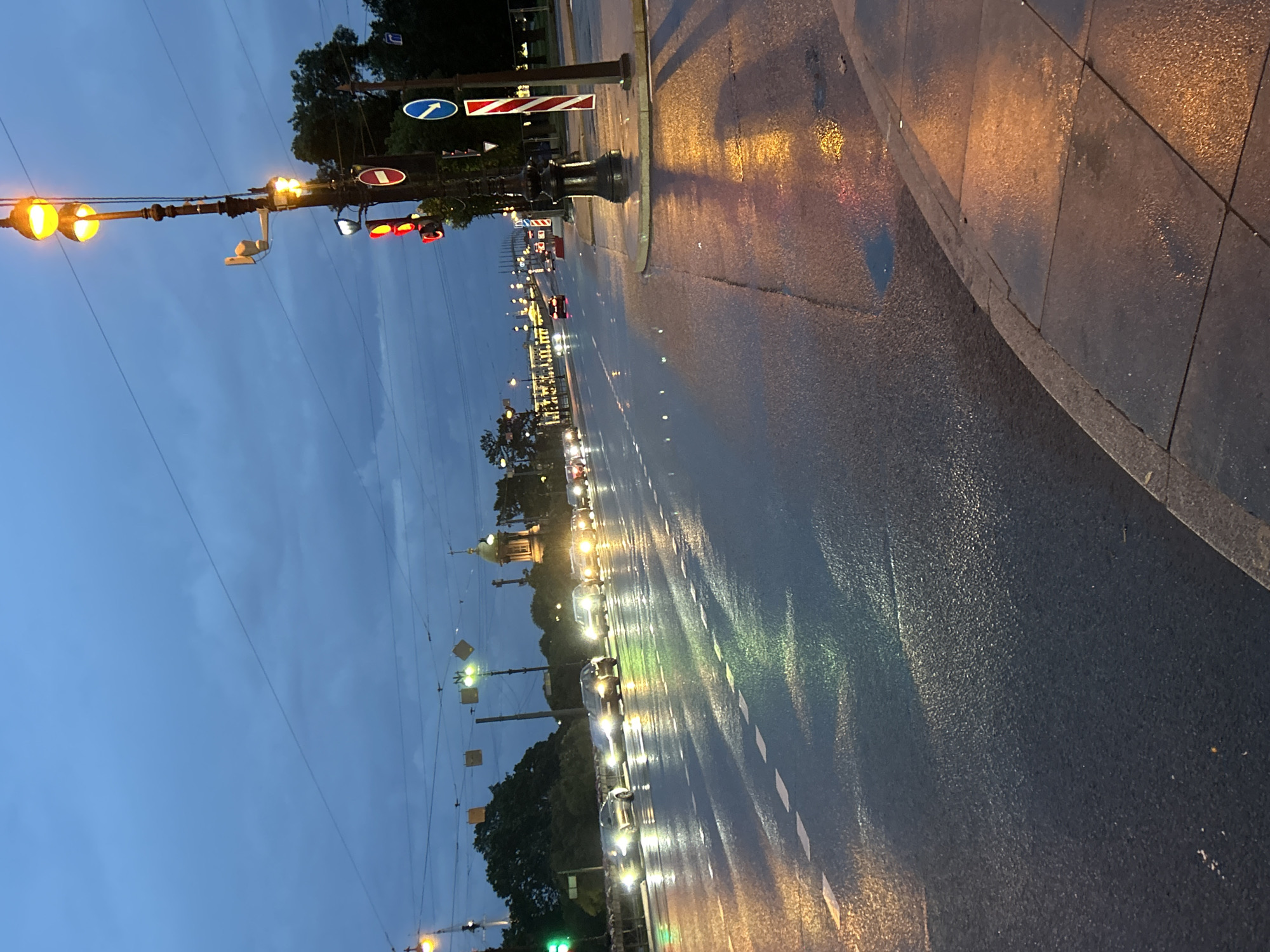 The image depicts a city street at dusk, likely after rain, with a wet asphalt road reflecting the lights of passing cars and street lamps. A line of cars is traveling in the distance, their headlights illuminated. A tall street lamp with multiple lights stands prominently on the right side of the image, accompanied by traffic signals and signs. Trees line the left side of the street, and in the far distance, a building with a dome is visible, suggesting a location with historical or significant architecture. The sky is a deep blue, with clouds indicating twilight or an overcast condition. The overall scene suggests a busy urban environment transitioning from day to night.