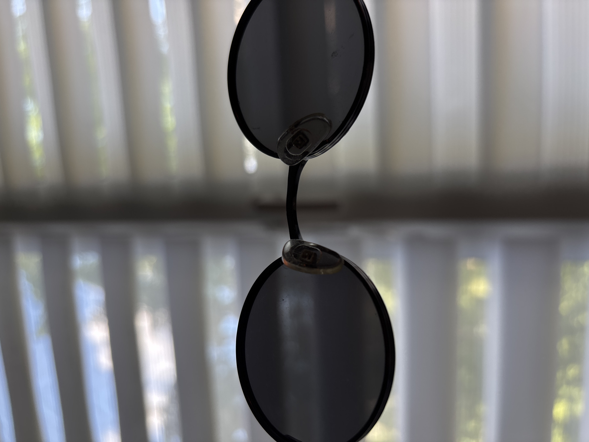 A pair of dark-rimmed oval sunglasses with tinted lenses is presented against a backdrop of white horizontal blinds. The blinds are partially open, allowing a blurred view of a bright, outdoor scene with streaks of blue sky and green foliage. The sunglasses are positioned so that one lens is slightly closer to the camera. There are no people or discernible interactions. The environment appears to be indoors, near a window. The image's origin in Saint Petersburg, Russia, cannot be determined from the visual information provided.