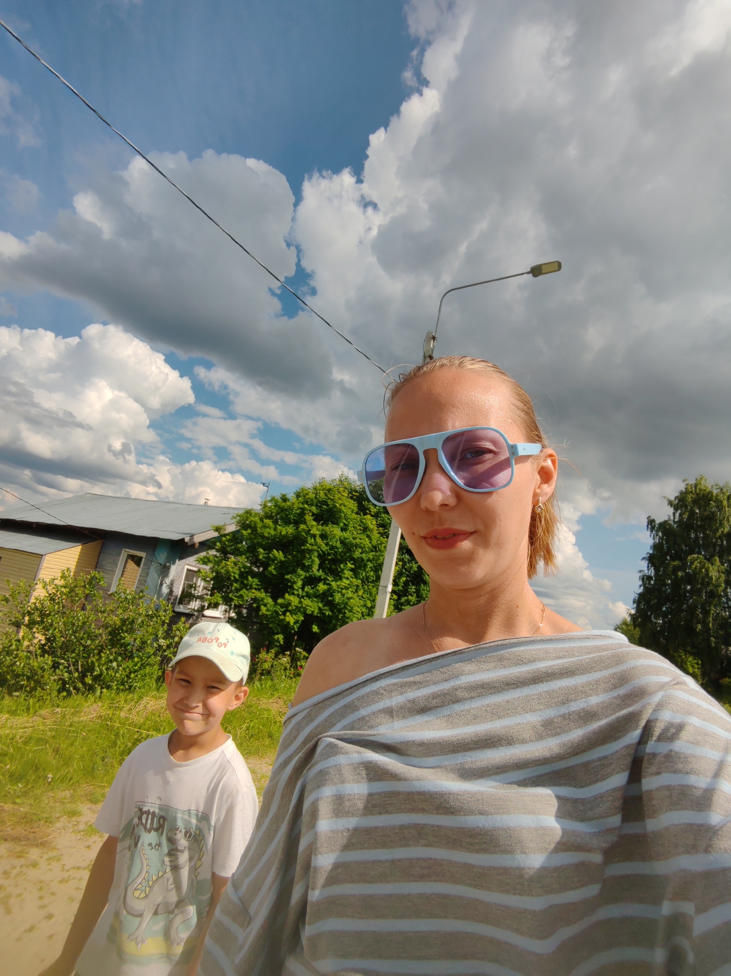 A woman wearing blue aviator sunglasses and a grey and blue striped off-the-shoulder top is taking a selfie outdoors. To her left and slightly behind her, a young boy wearing a white t-shirt with a dinosaur graphic and a light blue baseball cap is smiling. The background shows a house with a grey roof and yellow siding, green trees and bushes, a dirt path, and a cloudy sky with visible power lines and a street light. The image appears to be taken in a rural or suburban area in Чухлэм, Russia.