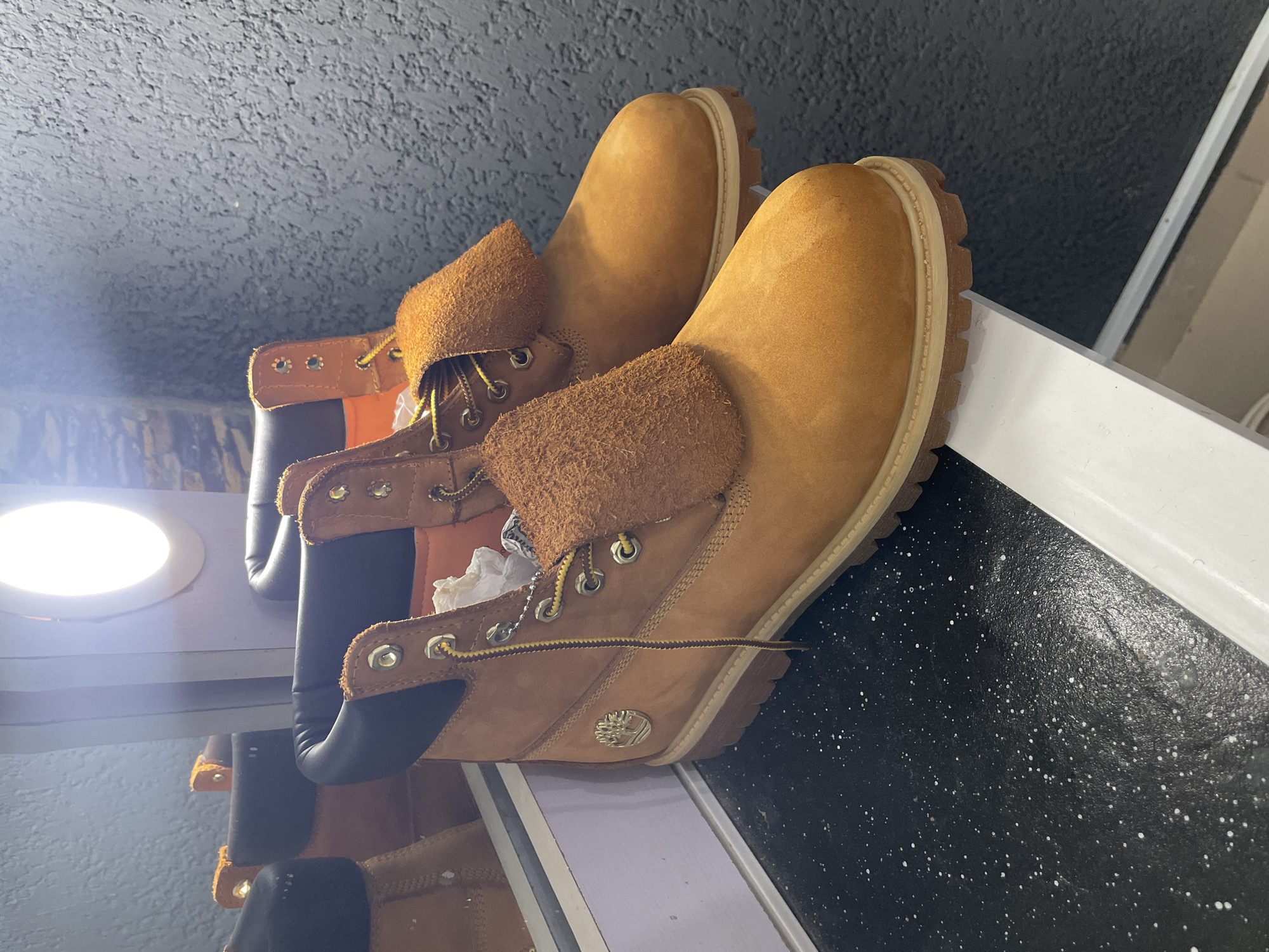 The image shows two Timberland boots placed on a surface that appears to be a black speckled floor next to a white shelf. The boots are the classic tan color with brown suede cuffs folded down. The lighting is bright, and some of the surrounding surfaces look to be reflective. There is not enough information to definitively determine if the image was taken in Jos, Nigeria.