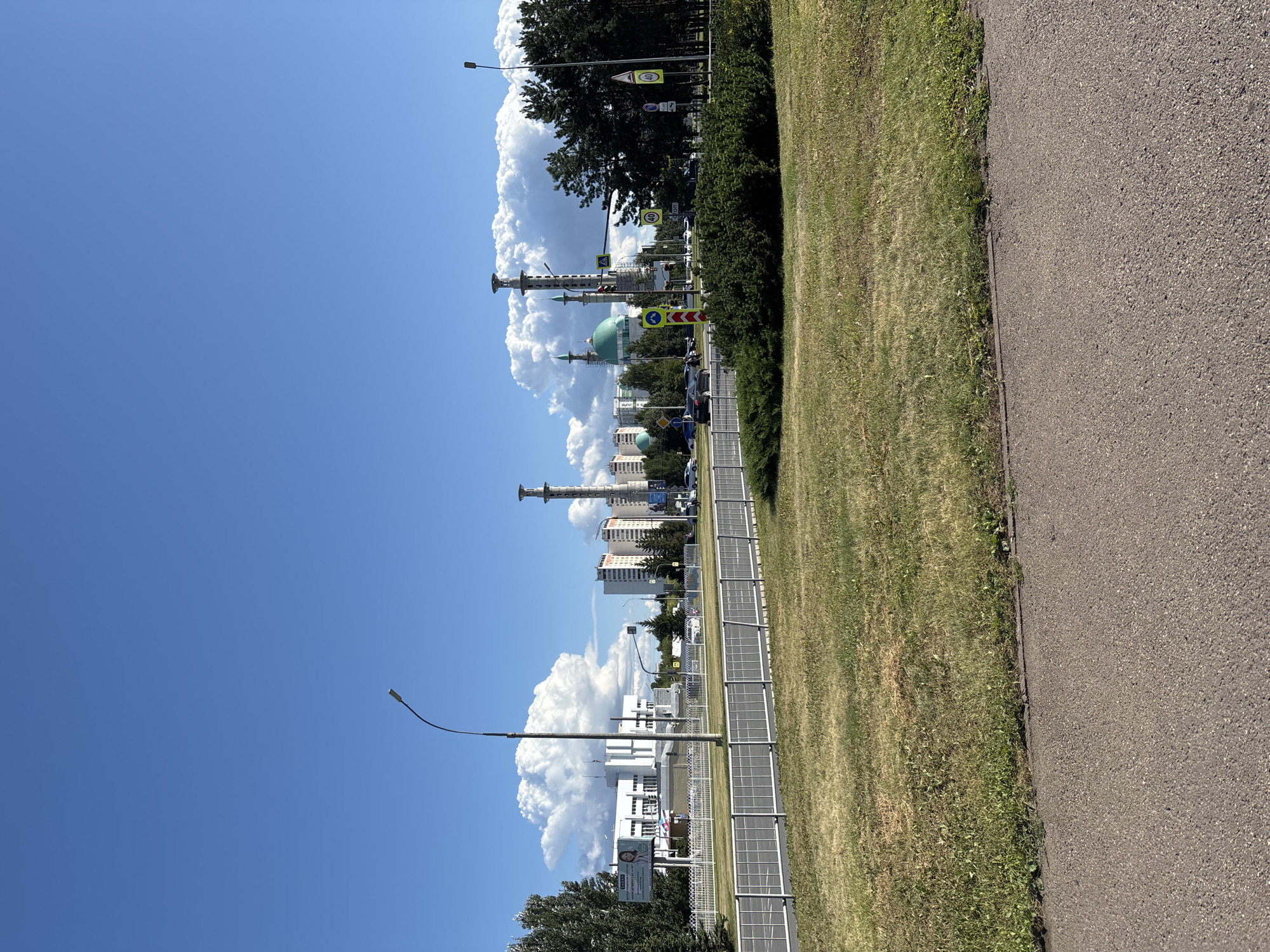 This is a medium-shot, eye-level, outdoor photograph taken on a sunny day in Naberezhnye Chelny, Russia. The foreground features a section of asphalt road, followed by a strip of dry, yellowing grass and a dense green bush. Behind the bush, a metal fence runs horizontally across the frame. Beyond the fence, a two-lane road is visible with several cars driving. A prominent white building with a large billboard advertising children's products is on the left, with a tall street lamp in front of it. Further into the background, the distinctive green domes and minarets of the Kamal Ad-Din al-Farisi Mosque are visible, flanked by tall, modern buildings. Several traffic signs, including speed limit signs, are present at an intersection. The sky is a clear, vibrant blue with large, puffy white clouds dominating the upper half of the image. The overall scene depicts an urban environment on a clear summer day, highlighting a blend of modern architecture and a prominent religious structure.