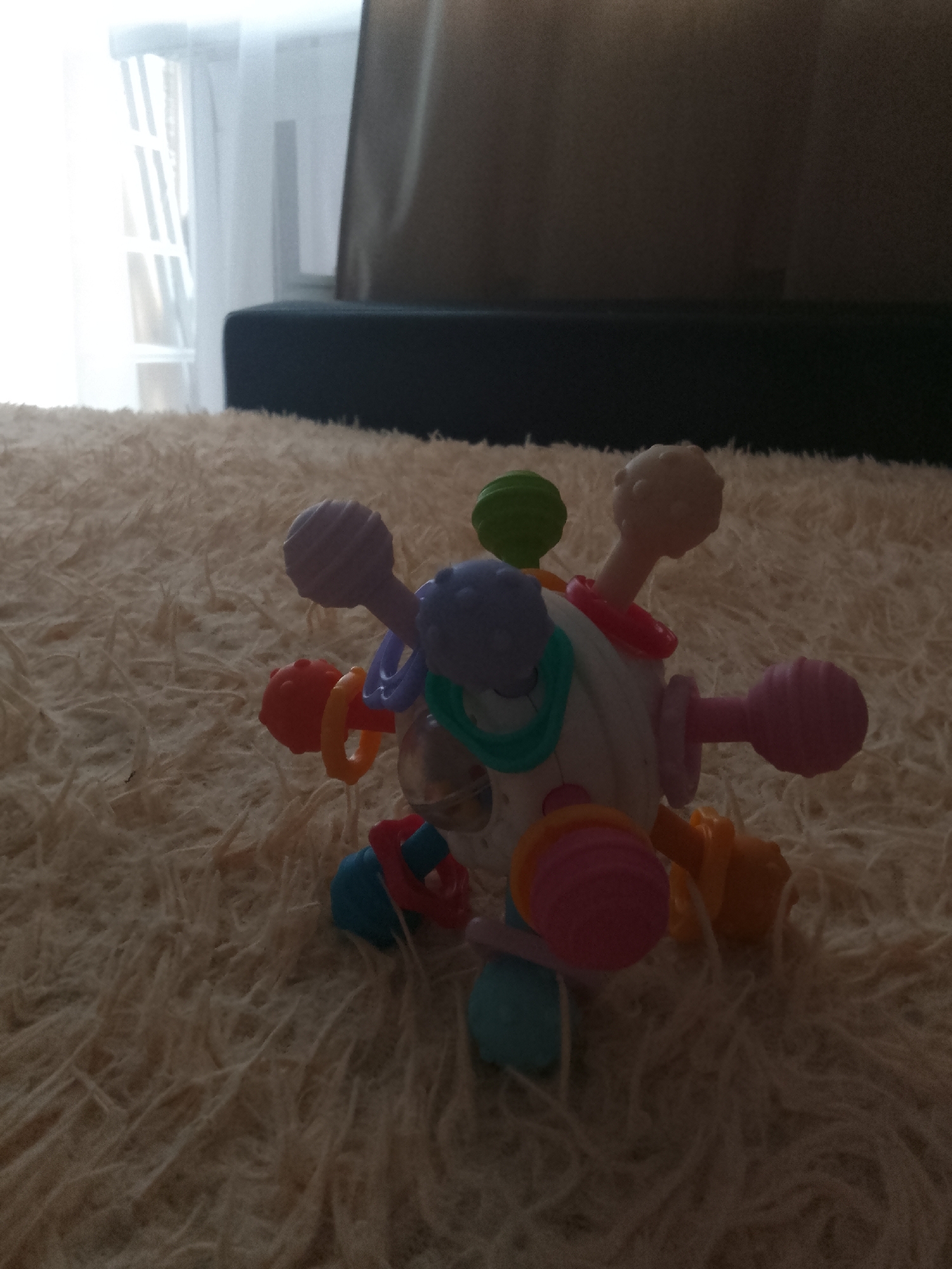 A colorful baby rattle is on a plush, light-colored carpet. The rattle is spherical with various colorful, textured appendages sticking out. The background is dark and out of focus, with what appears to be a window and curtains on the left side, and furniture on the right. There are no people present in the image.