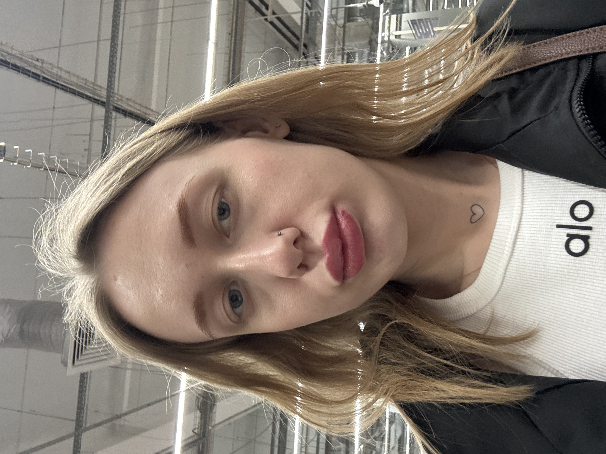 A close-up portrait shows a female individual centered, looking directly at the camera. She has blonde hair, blue eyes, and fair skin. A small silver stud is present in her left nostril piercing, and a clear stud earring is in her right earlobe. Her lips are coated with a dark pink cosmetic product; a small red abrasion or chapping is visible on her lower lip. A black outline heart tattoo is located on the right side of her neck, above the collarbone. She is wearing a white ribbed top with the black lowercase text "alo" printed on the chest, a thin gold chain necklace, and a black zippered jacket. A brown strap, possibly from a bag, is visible over her right shoulder.
The background depicts an indoor commercial or retail space characterized by a high, light-colored ceiling. Overhead, multiple horizontal fluorescent strip lights, metal structural beams, air conditioning vents, and a corrugated silver ventilation duct are visible. The lighting is bright and artificial. The specific location within Rostov-on-Don, Russia, cannot be determined from the visual information presented.