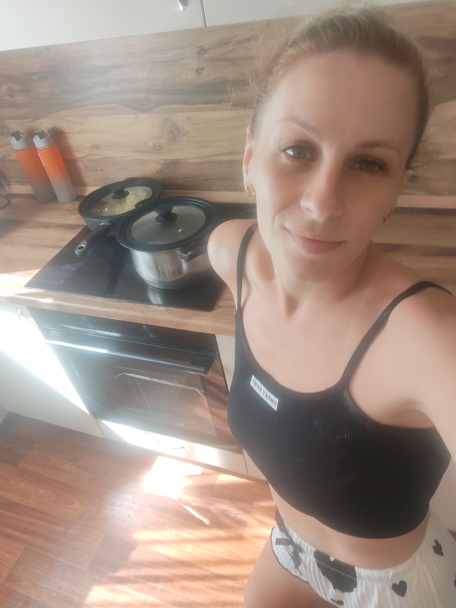 A woman with blonde hair pulled back is taking a selfie in a kitchen. She is wearing a black tank top with a white label and white patterned shorts. In the background, there are two pots on a stovetop, and two orange and gray bottles on a shelf. The kitchen has wooden cabinets and a wooden countertop. Sunlight streams into the room from the right, casting bright reflections on the stovetop and floor.