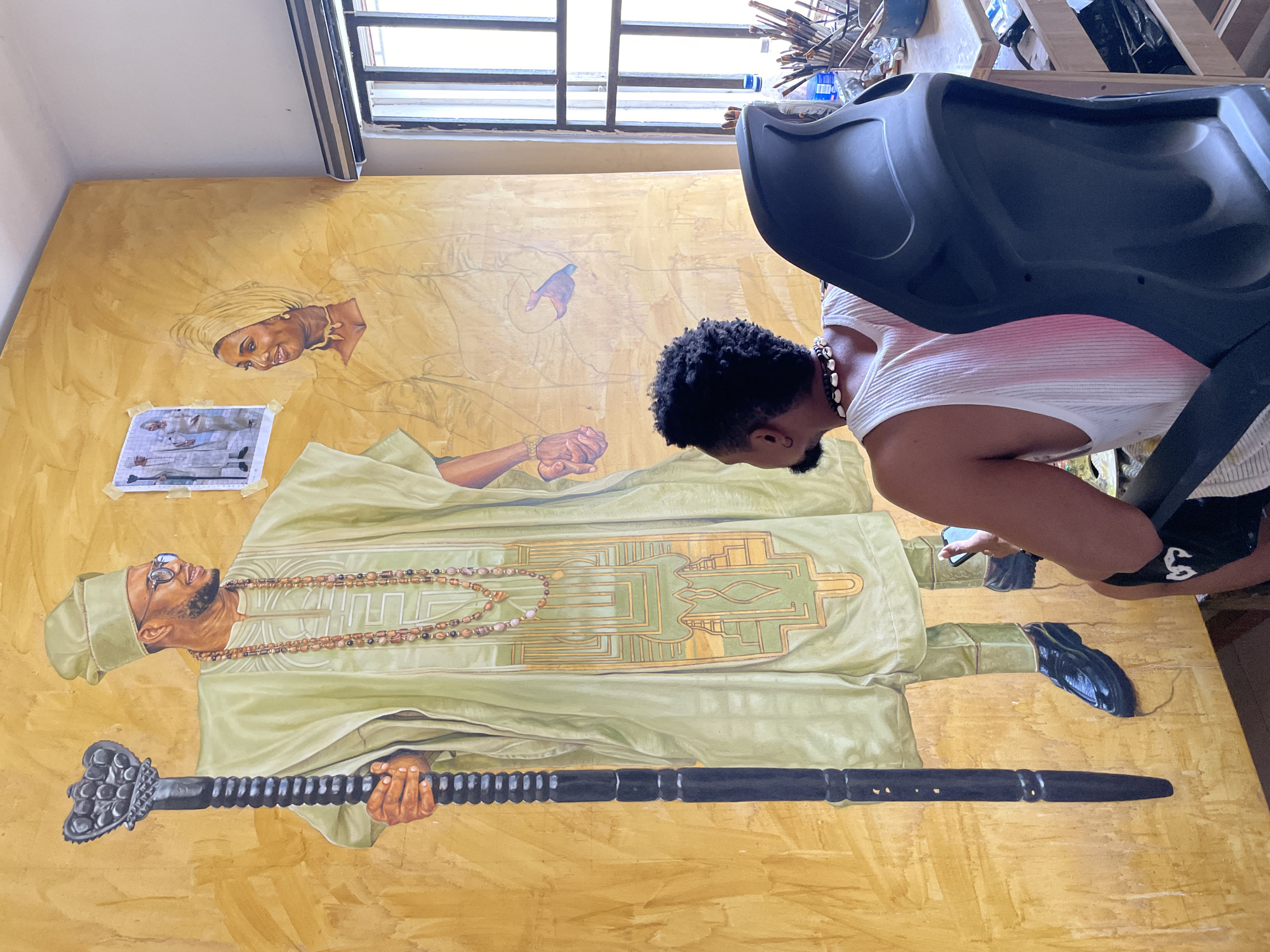An artist is in their studio in Ado, Nigeria, working on a large mural. The mural depicts a man and a woman in traditional attire. The man, on the left, is wearing a light green agbada and holding a dark, ornate staff. He has glasses and a beard. The woman, on the right, is wearing a matching light green outfit and headwrap, with a necklace. A reference photo of two people in similar attire is taped to the wall above and behind the mural. The artist, a Black man with curly hair and a beard, is seated in a black chair, viewed from behind and the side, looking at his phone. He is wearing a white tank top and a shell necklace. The studio appears to be well-lit with natural light coming from a window on the right, which has vertical bars. A table with art supplies, including brushes and bottles, is visible near the window. The wall behind the mural has a textured, golden finish.