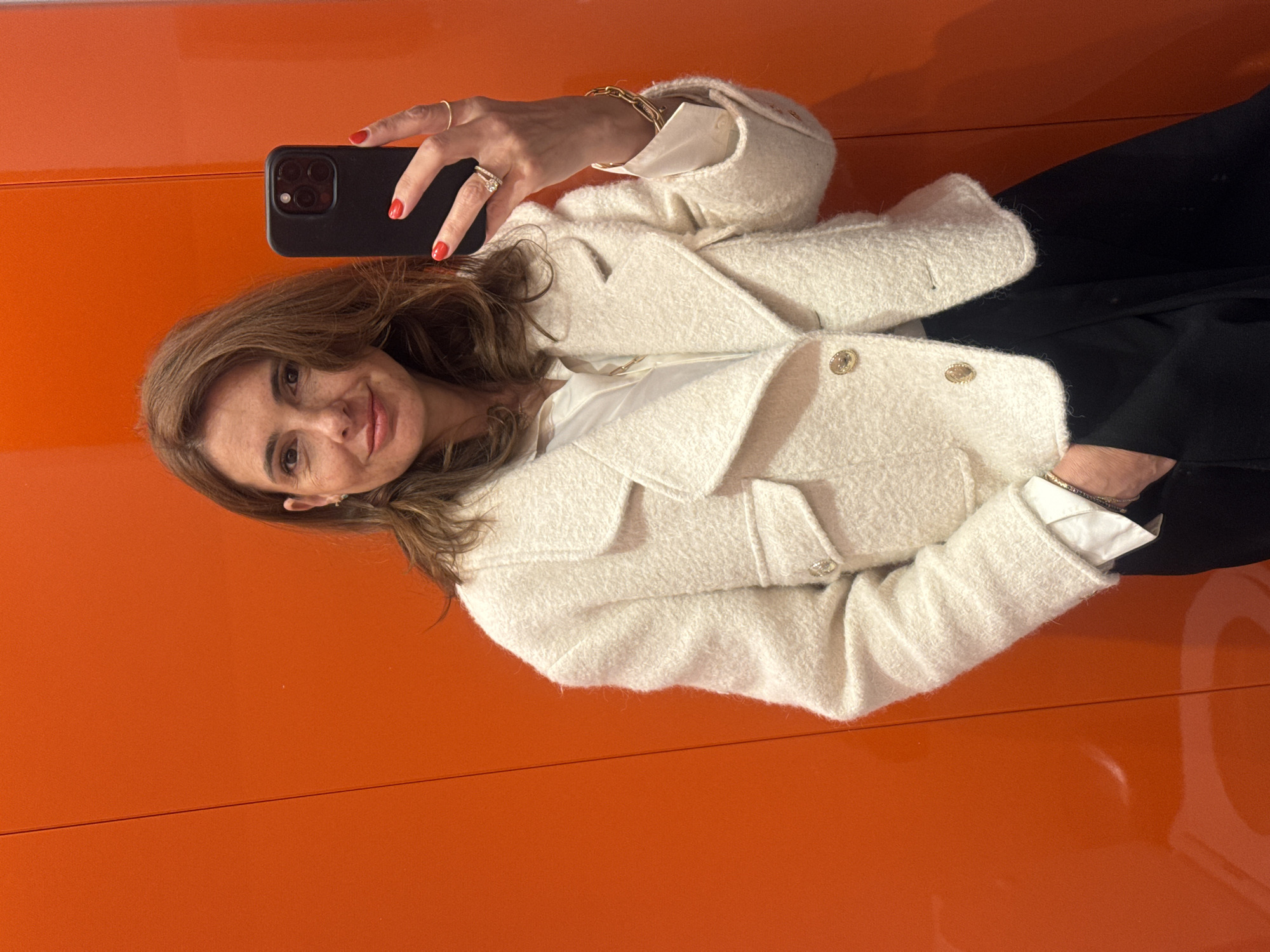 A woman in a white jacket and black pants is holding a cell phone while standing in front of an orange wall or object. The location in Lisbon, Portugal is not evident from the image.