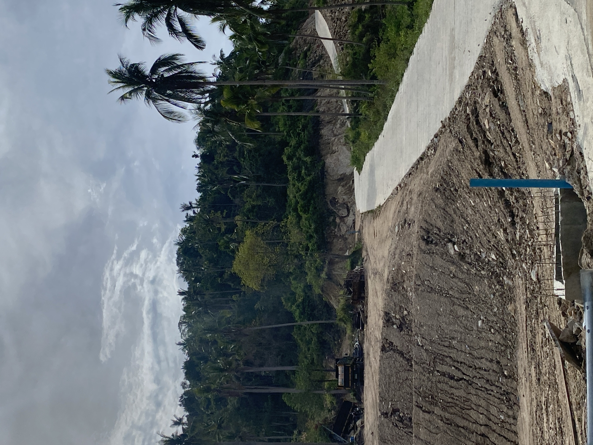 The image depicts an outdoor scene, possibly road construction, in the vicinity of Baan Wang Ta Kien, Thailand. The sky is overcast with white clouds. A hillside covered in dense tropical vegetation, including numerous tall palm trees, rises on the left side of the frame. A partially constructed road or retaining wall runs along the right side of the image, with exposed dirt and gravel. Construction materials and machinery are visible near the base of the hillside, suggesting ongoing construction activity. A blue object is seen on the edge of the road.