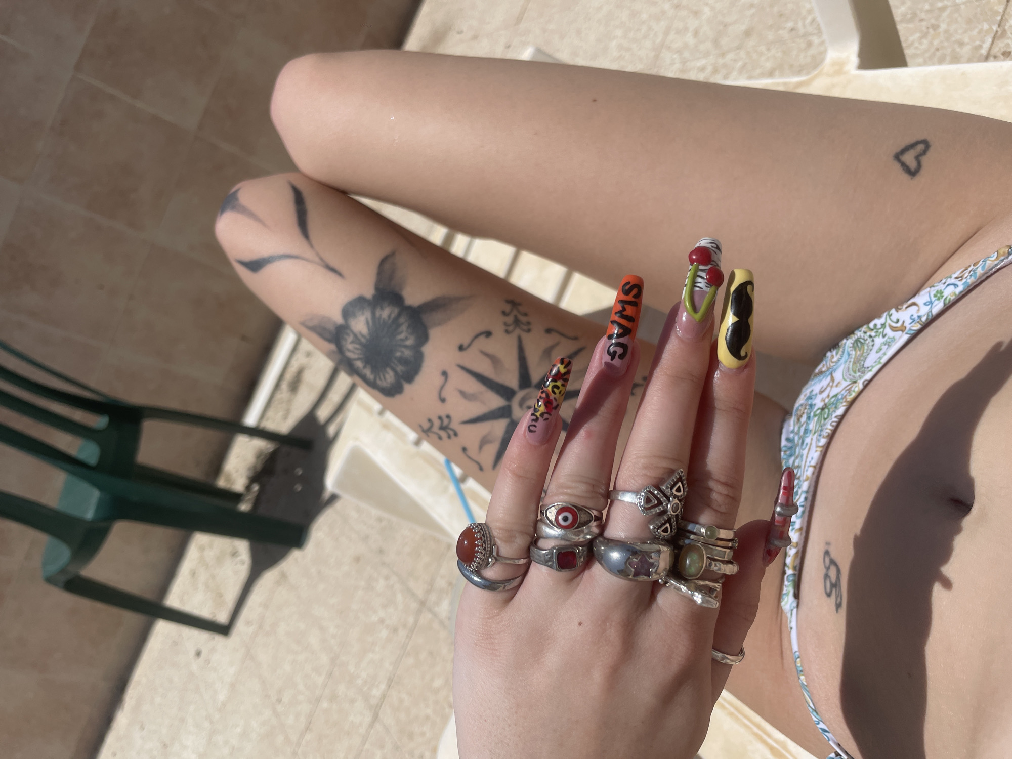 A person is reclining outdoors, likely in a pool or sunbathing area. The setting appears to be in a warm, sunny location. The foreground is dominated by the person's hand, heavily adorned with multiple rings on each finger, including rings with stones, silver bands, and unique designs such as a butterfly and an eye. The person has long, elaborately designed fingernails, with different patterns and words like "SWAG" on them.
The person is wearing a patterned bikini top. Tattoos are visible on the person's body, including a heart near the upper chest, a flower and compass design on the arm, and another small tattoo near the side of the torso.
The background shows tiled flooring and a green plastic lawn chair. The scene is bathed in bright sunlight, creating sharp shadows.
There is no evidence that this image was taken in Petah Tikva, Israel.