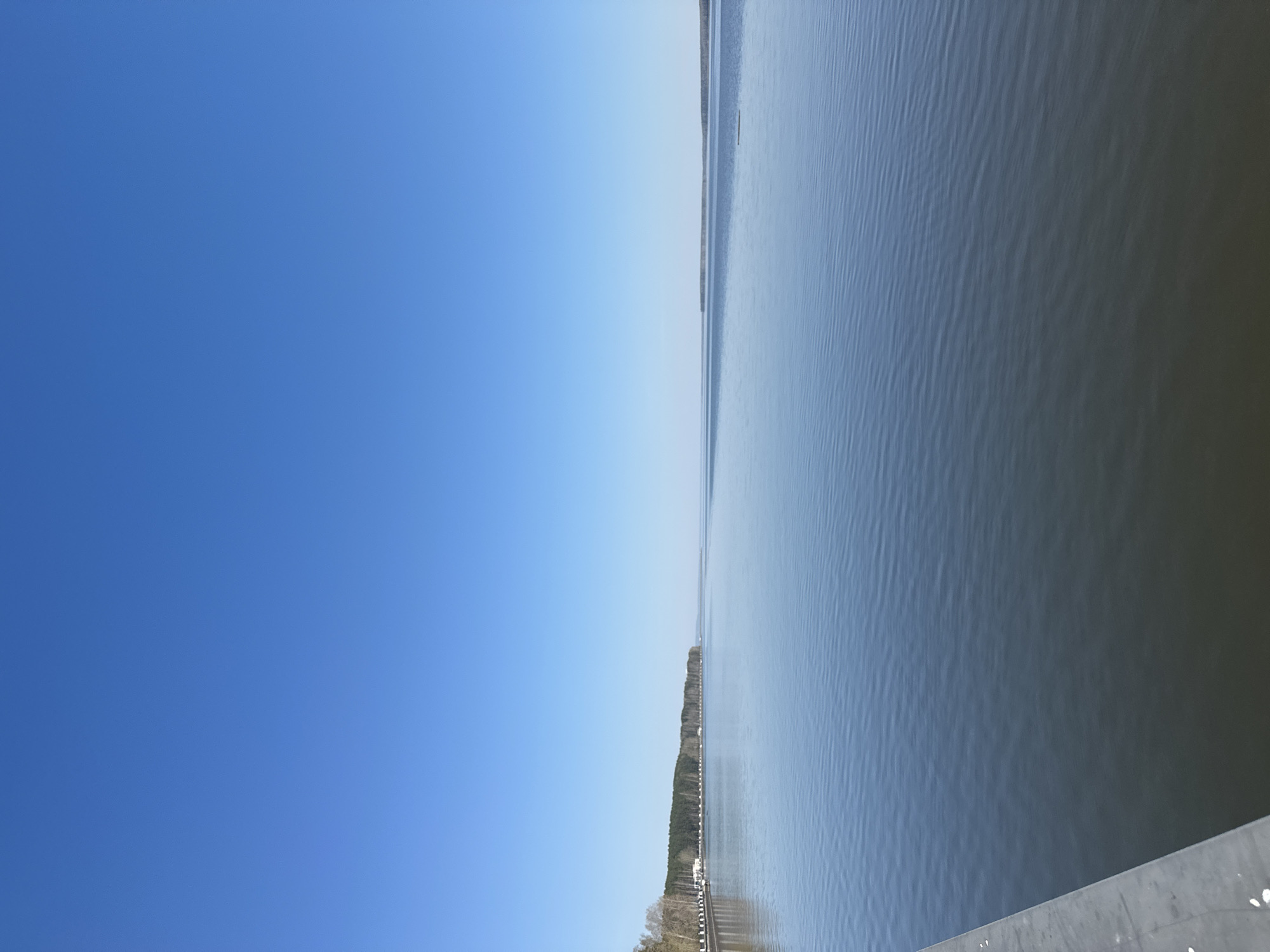 The image shows a horizontal view of a body of water, likely a river or lake, with a clear blue sky above. A strip of land with trees and possibly buildings is visible on the far side of the water. The water surface shows slight ripples, suggesting a gentle breeze. The location is likely Chaikovsky, Russia, given the request.
