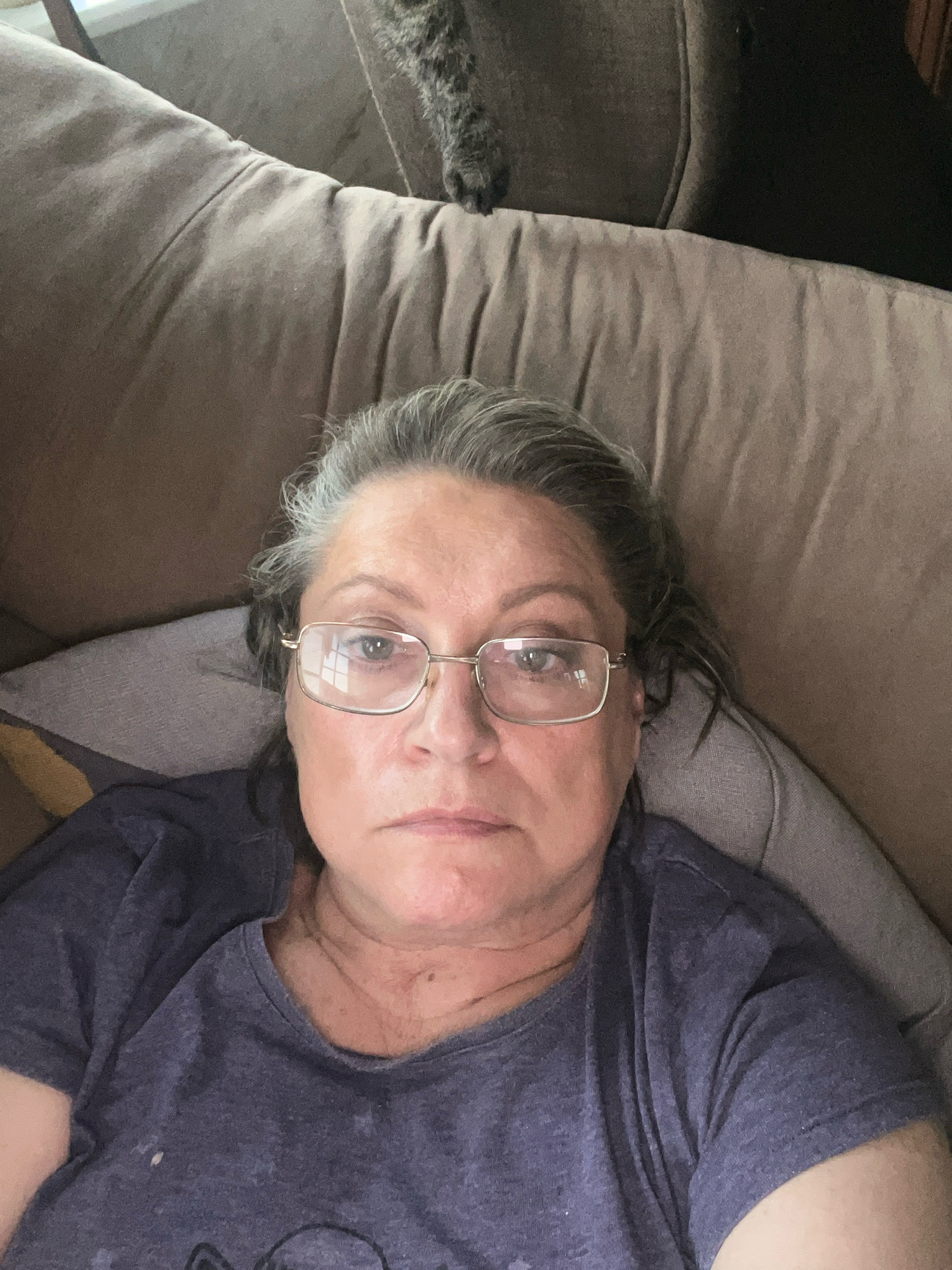 A selfie shows a middle-aged woman with grey hair and glasses resting on a brown couch. She is wearing a dark blue t-shirt. The background is the brown couch and a hint of dark fur from a pet. The image is taken from a high angle, looking down at the woman. The lighting is dim, creating a shadow on the right side of her face. There is no discernible location in the city of Vladimirovka, Russia.