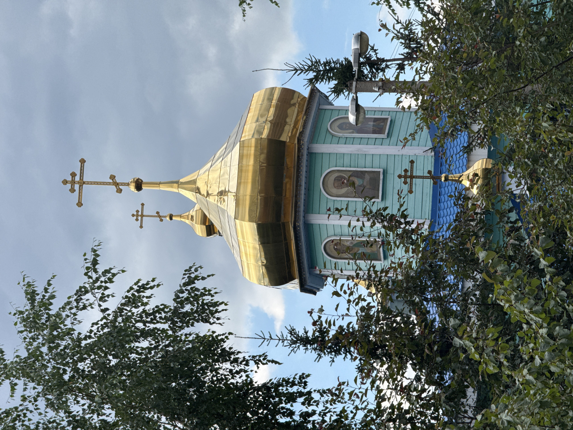 An exterior, eye-level, medium shot captures the upper section of a light blue church with a bright gold dome, topped by a gold, multi-tiered cross. The gold dome is octagonal and reflective, catching the light of a cloudy sky. Two arched windows with religious iconography are visible on the church wall below the dome. Trees with green leaves partially obscure the view of the church, with a thin evergreen tree also visible on the right side of the frame. A utility pole with two streetlights is positioned to the right of the church. The roofline beneath the gold dome is painted blue. The scene is set against a backdrop of a cloudy sky with patches of blue peeking through.