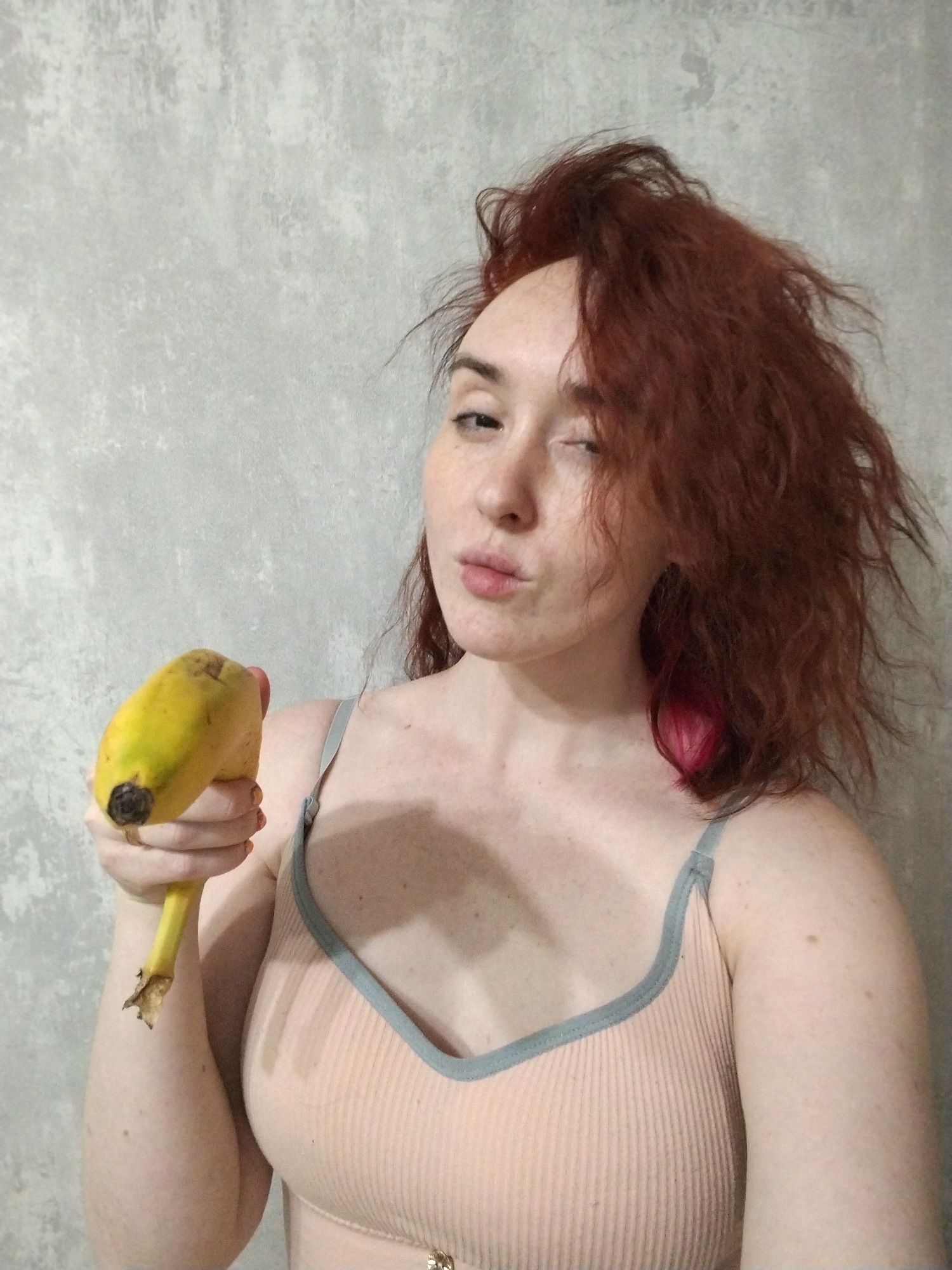A female individual is depicted from the upper chest upwards, positioned slightly right of the frame's center. She has reddish-brown, disheveled hair, with a distinct streak of bright pink hair visible on her right side near the shoulder. Her left eye is partially closed, while her right eye is open, and her lips are pursed. She wears a ribbed, light peach-colored spaghetti-strap top with light blue trim. Her left hand, with fingernails painted orange, holds a single ripe yellow banana horizontally, presenting it towards the camera. The banana exhibits minor brown spots and a detached stem end. The background is a uniform light grey wall with irregular surface texture. The specific location within the city of Izhevsk, Russia, cannot be determined from the visual information provided.