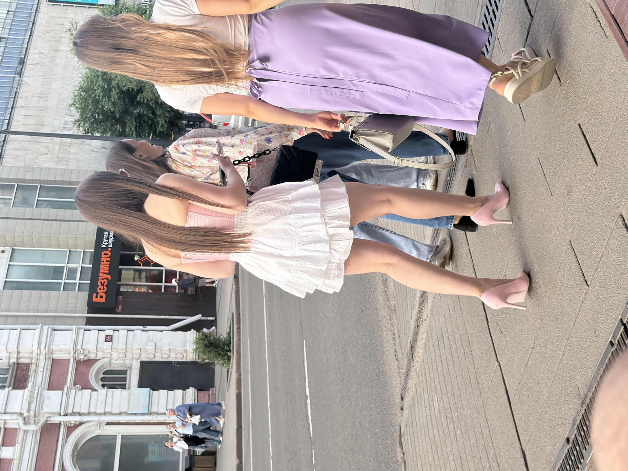 The image shows a street scene in Krasnoyarsk, Russia. In the foreground, three young women are walking on a sidewalk. The woman on the left is wearing a pink strapless top and a short white ruffled skirt, with pink high heels. The woman in the middle is wearing a floral print blouse and blue jeans. The woman on the right is wearing a white t-shirt and a long, flowing lavender skirt, with beige sandals. They are walking away from the camera. In the background, there is a building with red and white architectural details and a sign that reads "Безумно. Крутая шаурма" which translates to "Crazy. Cool Shawarma". There are other people walking on the sidewalk and across the street. The environment appears to be urban, with a paved road and sidewalk.