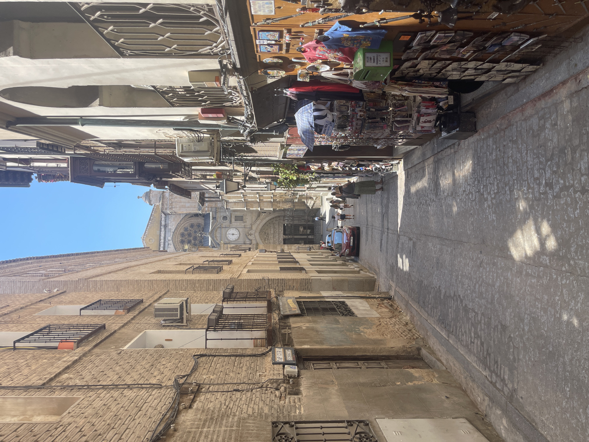 This image captures a narrow, cobblestone street in Toledo, Spain, likely in the historic old town. On the left, a tall, brick building with multiple windows and balconies lines the street. An air conditioning unit is mounted on the wall of this building. A sign reading "TAPICERIA DECORACION" is visible above a doorway.
On the right side of the street, a different building features a series of arched openings, some of which are enclosed with ornate metal railings. This building houses a shop with an awning displaying various goods, including clothing, bags, umbrellas, and postcards. Several people are browsing or walking down the street.
Further down the street, a red car is parked. In the distance, the facade of a grand cathedral, likely the Toledo Cathedral, is visible with its clock tower. The sky is clear and blue, indicating a sunny day. The overall scene suggests a bustling, old-world European street with shops and historical architecture.