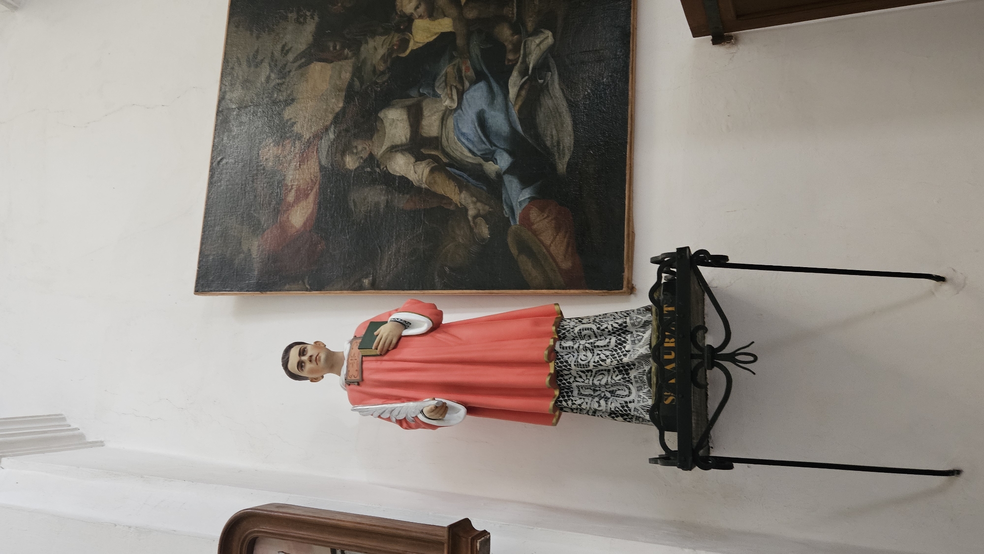 The image depicts a statue of a male figure in a red robe and a black and white patterned skirt, holding a book and a feather. The statue is positioned on a black metal stand with intricate detailing, and a plaque that reads "ST. LAURENT". To the right of the statue is a large painting depicting several figures, including a woman in a blue garment and an infant. The background is a plain white wall, with a decorative molding at the top left and a wooden door frame visible on the right. The scene appears to be indoors, possibly in a church or a museum, given the religious imagery and artwork. The statue and painting are illuminated by ambient light. The overall impression is one of religious art and veneration. This scene is located in the Église Saint-Antoine in Sant'Antonino, France.
