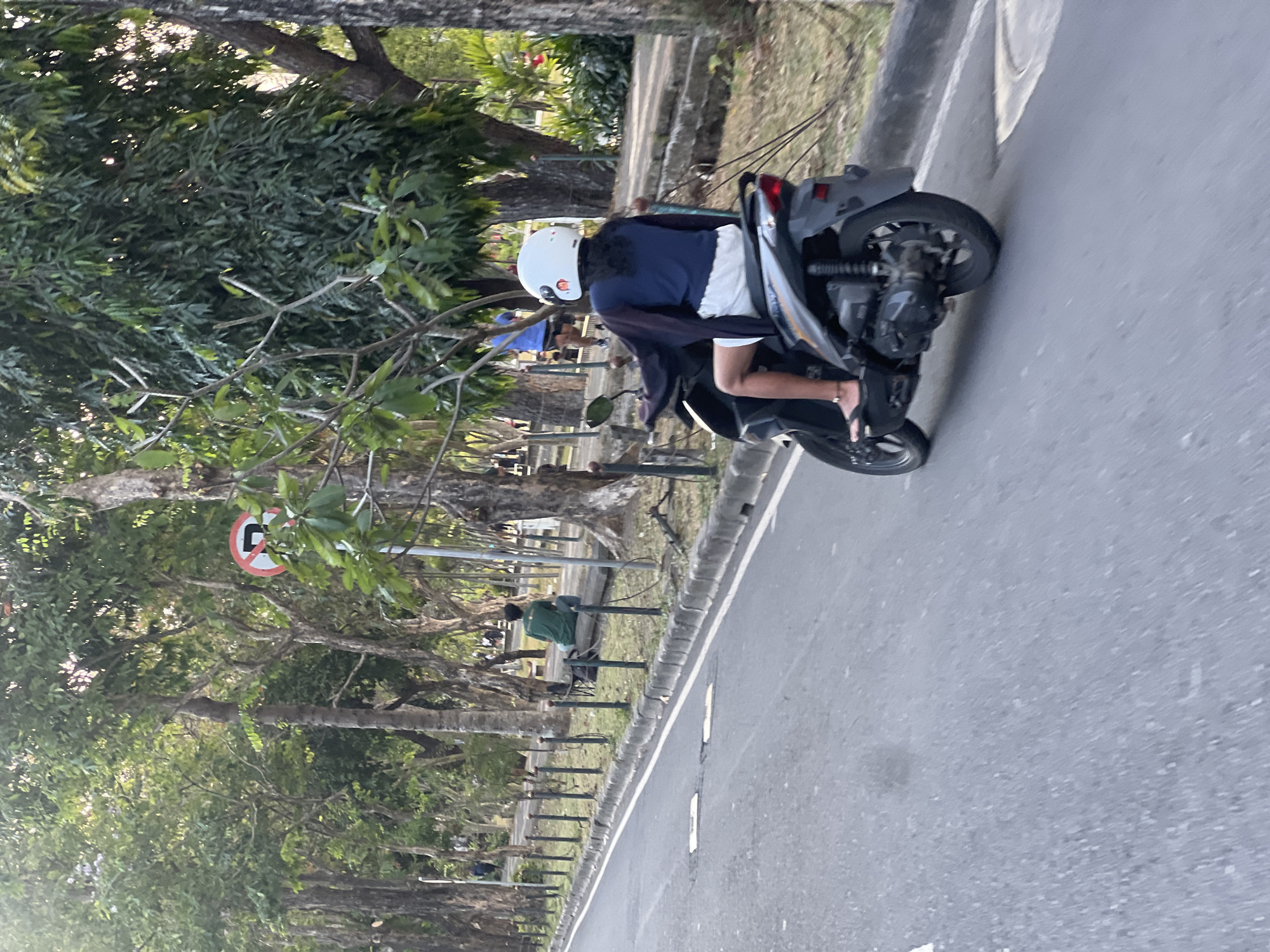 The image captures a street scene in Denpasar, Indonesia, likely during daytime given the lighting. In the foreground, a person is riding a scooter. The rider is wearing a white helmet, a dark blue shirt, and white shorts. Their legs are visible, and they are wearing flip-flops. The scooter is dark gray.
The road is paved and appears to be a street or boulevard with trees lining the side. A white line marks the edge of the road. Along the side of the road, there is a pathway or sidewalk, separated from the road by a low fence. Beyond the fence, people are seated on a long log, and other individuals are visible in the background, some walking. There are numerous trees with dense foliage, creating a shaded environment. A traffic sign, circular with a red border and a horizontal bar, is visible among the trees on the left side of the frame. The overall environment suggests a public space within the city, possibly a park or a tree-lined avenue.