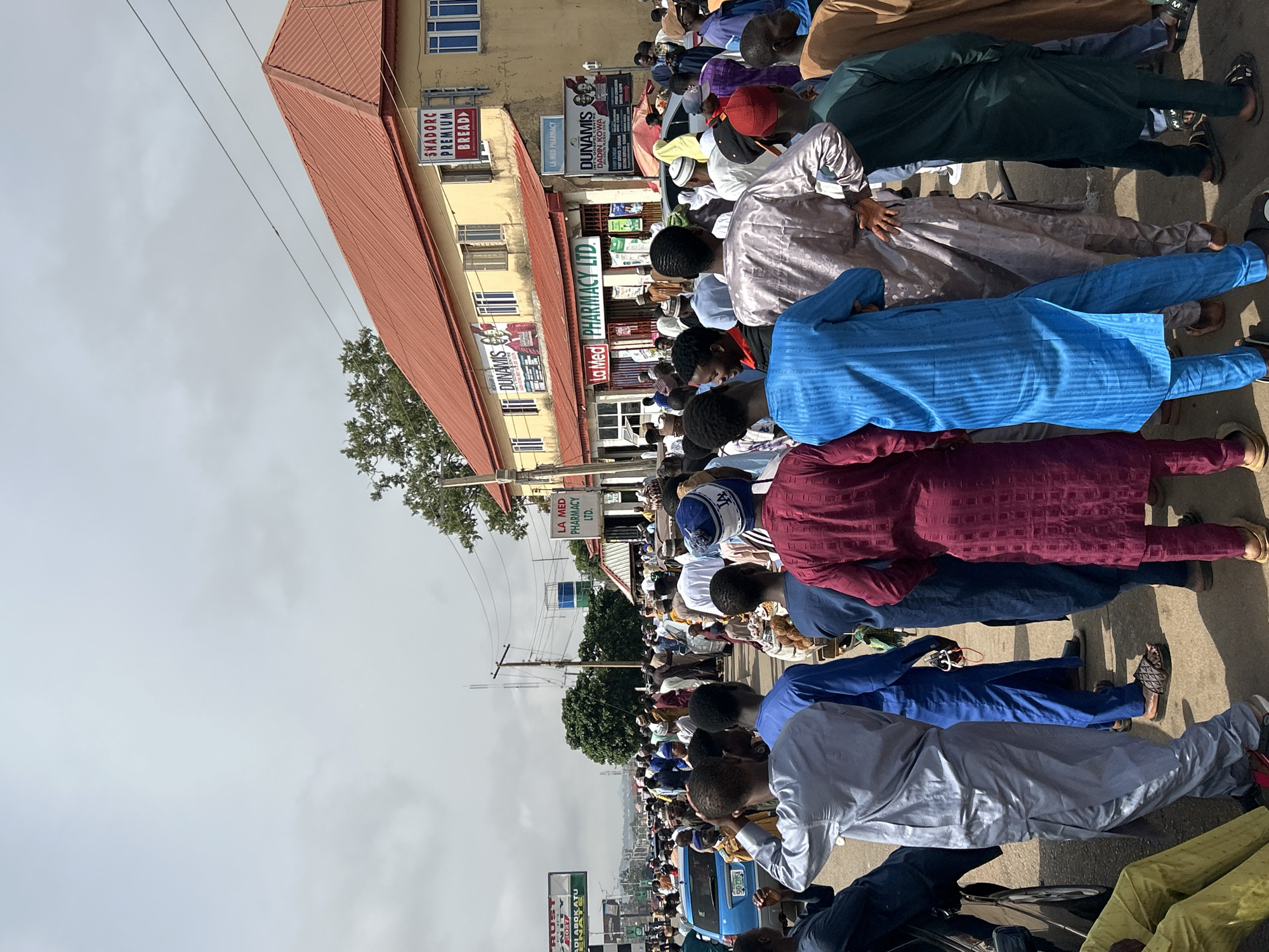 A large group of people are arranged in rows, facing the same direction, seemingly gathered on a street in Jos, Nigeria. Many are wearing traditional clothing, particularly long, flowing robes in various colors such as blue, maroon, and silver. Some individuals are wearing caps or head coverings. The street is partially blocked by the crowd. Buildings are visible in the background, with commercial signs displaying names like "SHADORC PREMIUM BREAD," "DUNAMIS," and "LA MED PHARMACY LTD." A blue vehicle is parked on the side of the street. Utility poles and wires are present above the street. The sky is overcast. A street sign appears to read "DIABOKATU OKATU".