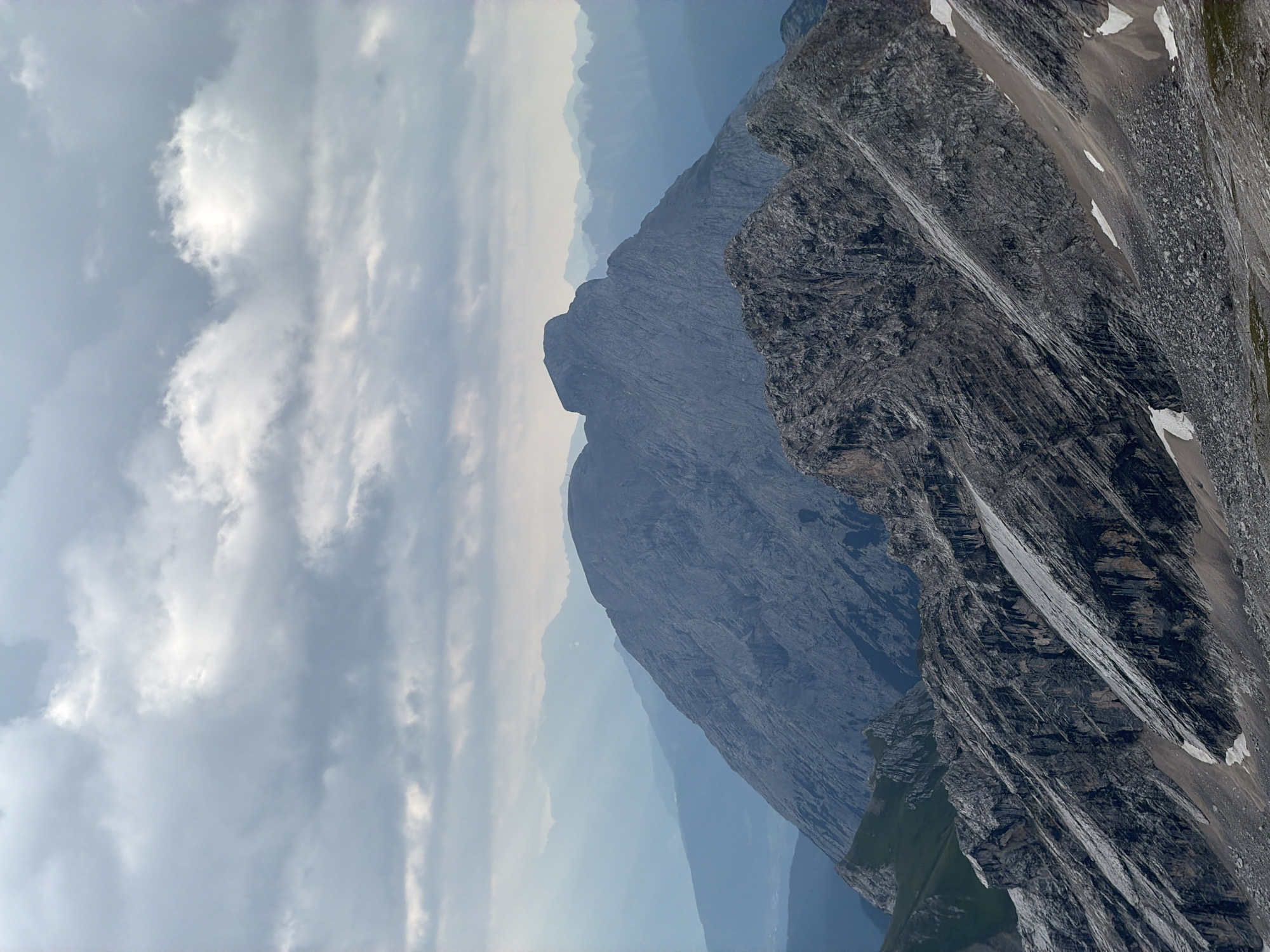 The image captures a panoramic view of jagged, rocky mountain peaks under a dramatic, overcast sky. The foreground features a steep, scree-covered slope with patches of snow. Behind it, a large, imposing mountain with a distinctive, somewhat flat summit dominates the mid-ground, its rocky face showing deep fissures and striations. Further in the distance, layers of muted blue mountain ranges recede into a hazy horizon. The overall impression is one of rugged, natural grandeur and a wild, untamed landscape. This scene is characteristic of the Bavarian Alps, a region that includes Garmisch-Partenkirchen, Germany.